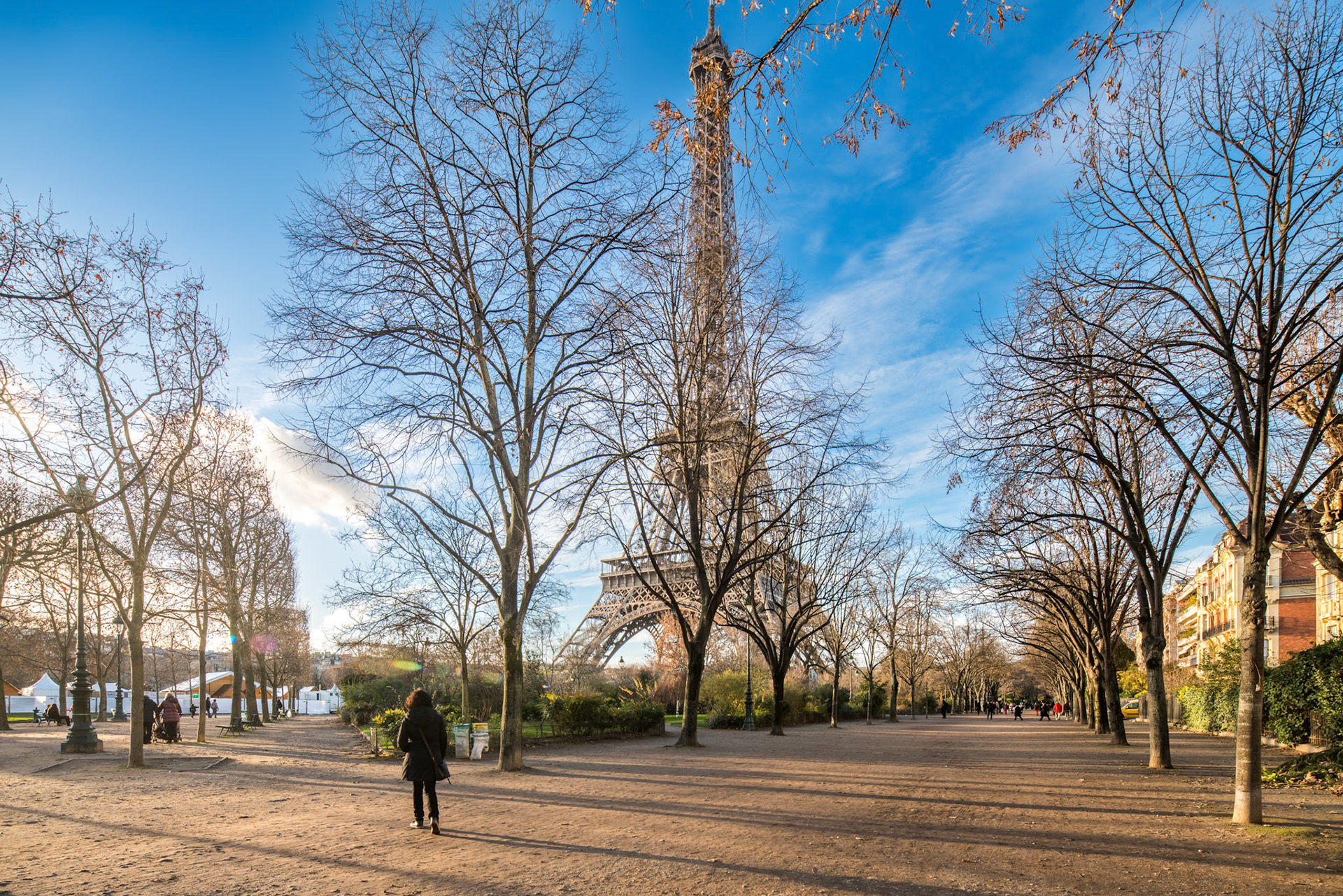 The Eiffel Tower rises above leafless trees as people stroll through a Paris garden at twilight.