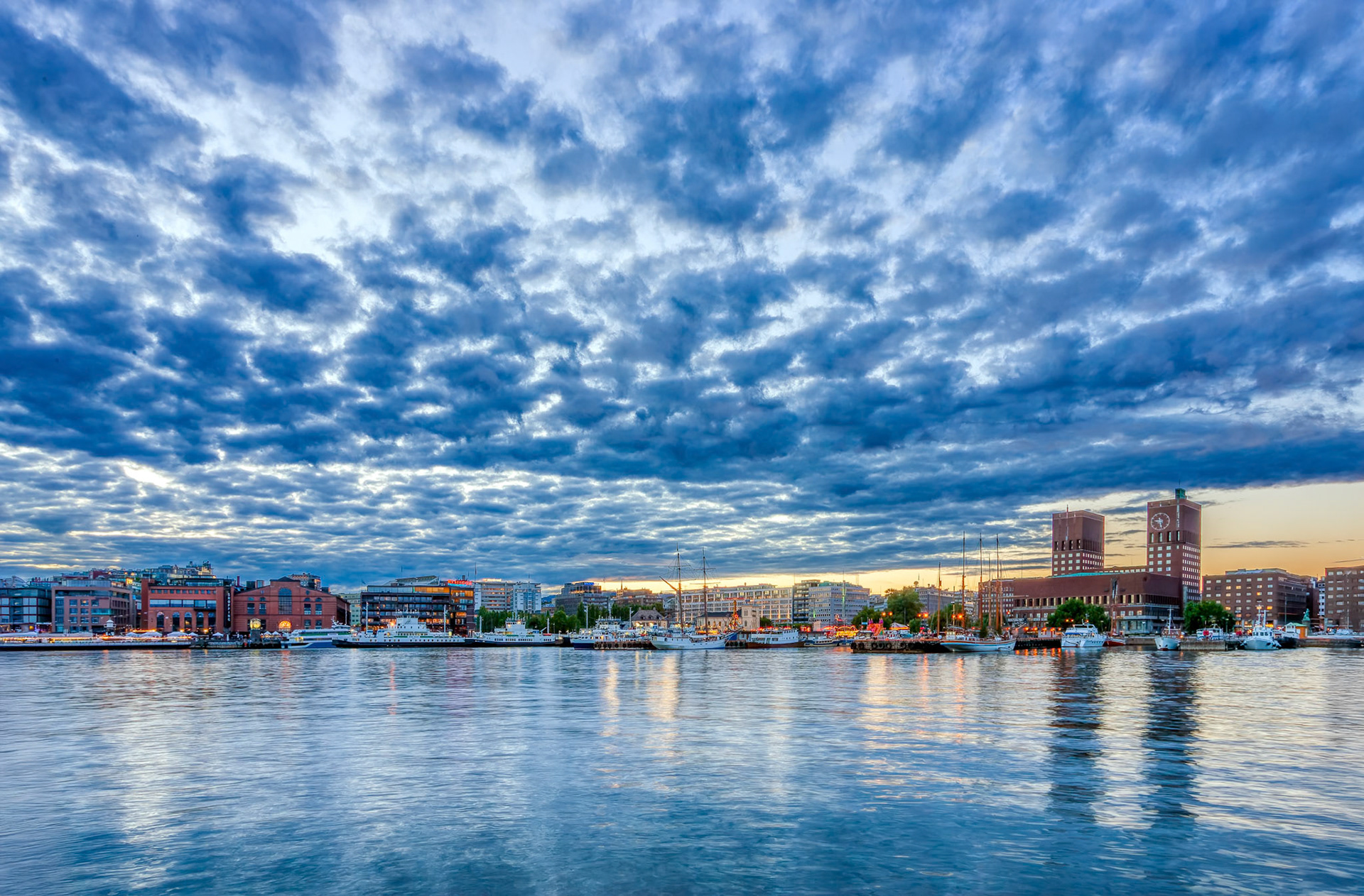 Twilight settles over Oslo, casting a warm glow on City Hall and Aker Brygge, with gentle waves reflecting the vibrant sky.