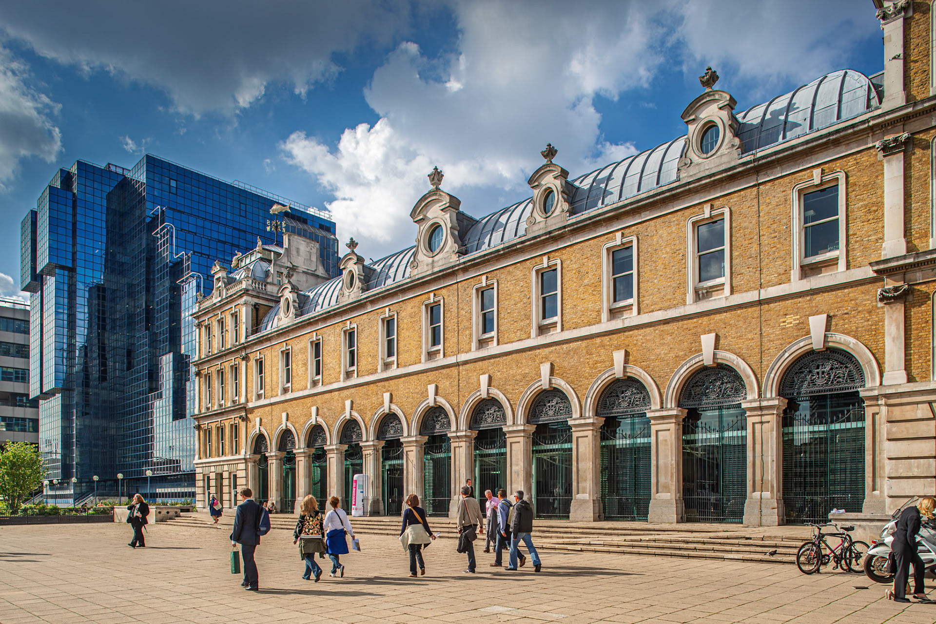 People walk past the Old Billingsgate Market building while the Northern and Shell Building stands nearby on the river bank.
