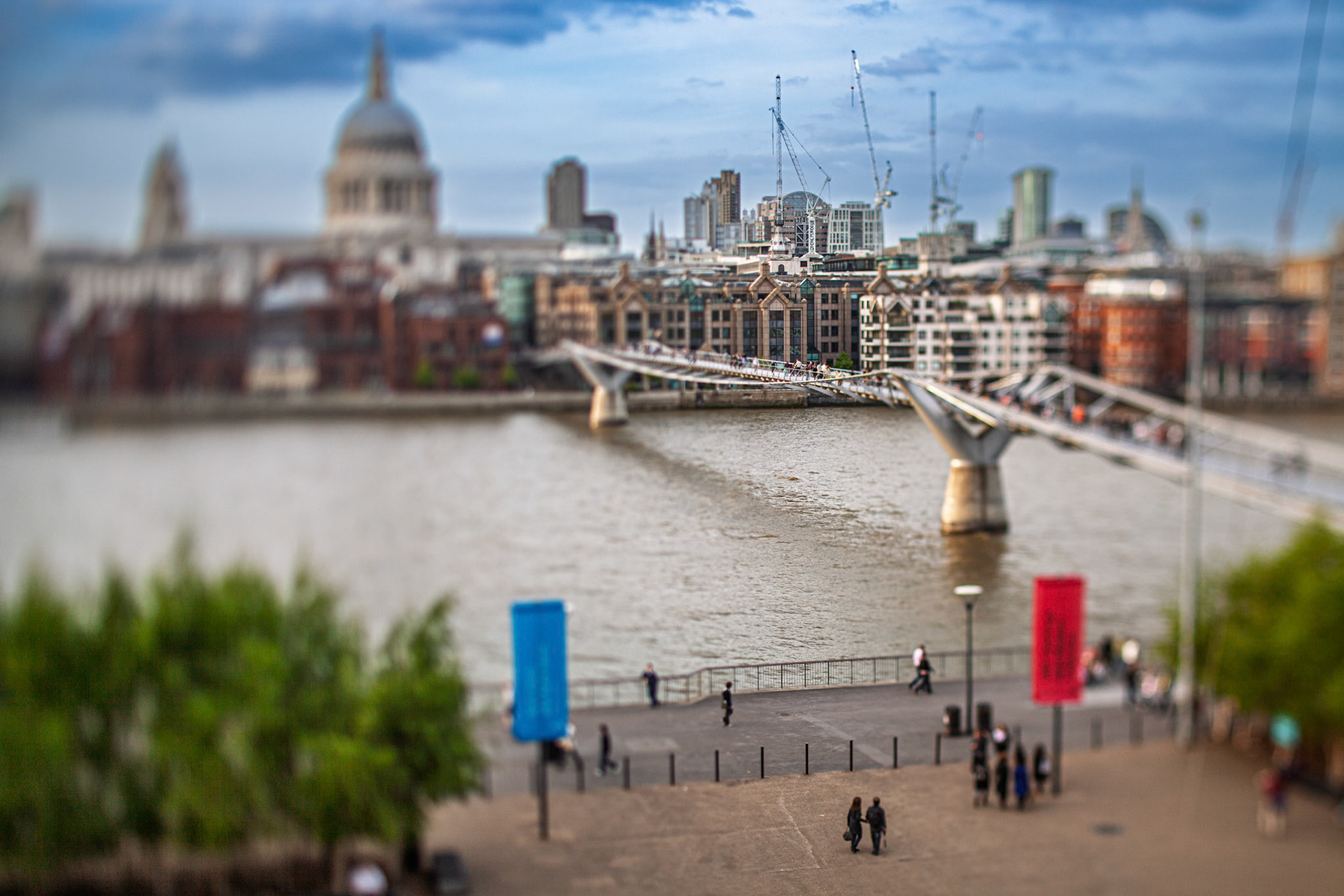 Millenium Bridge stretches over the Thames River, with St Paul Cathedral in the background. Visitors walk along the riverside in London, UK.