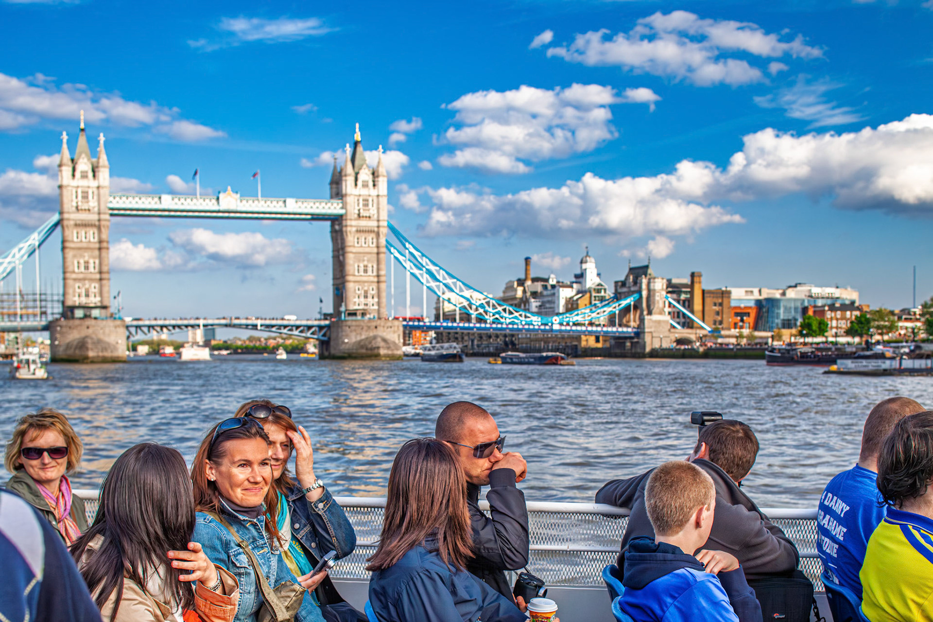 A group of tourists takes a boat ride on the river with Tower Bridge in view. They smile and point as they enjoy the city sights in London.