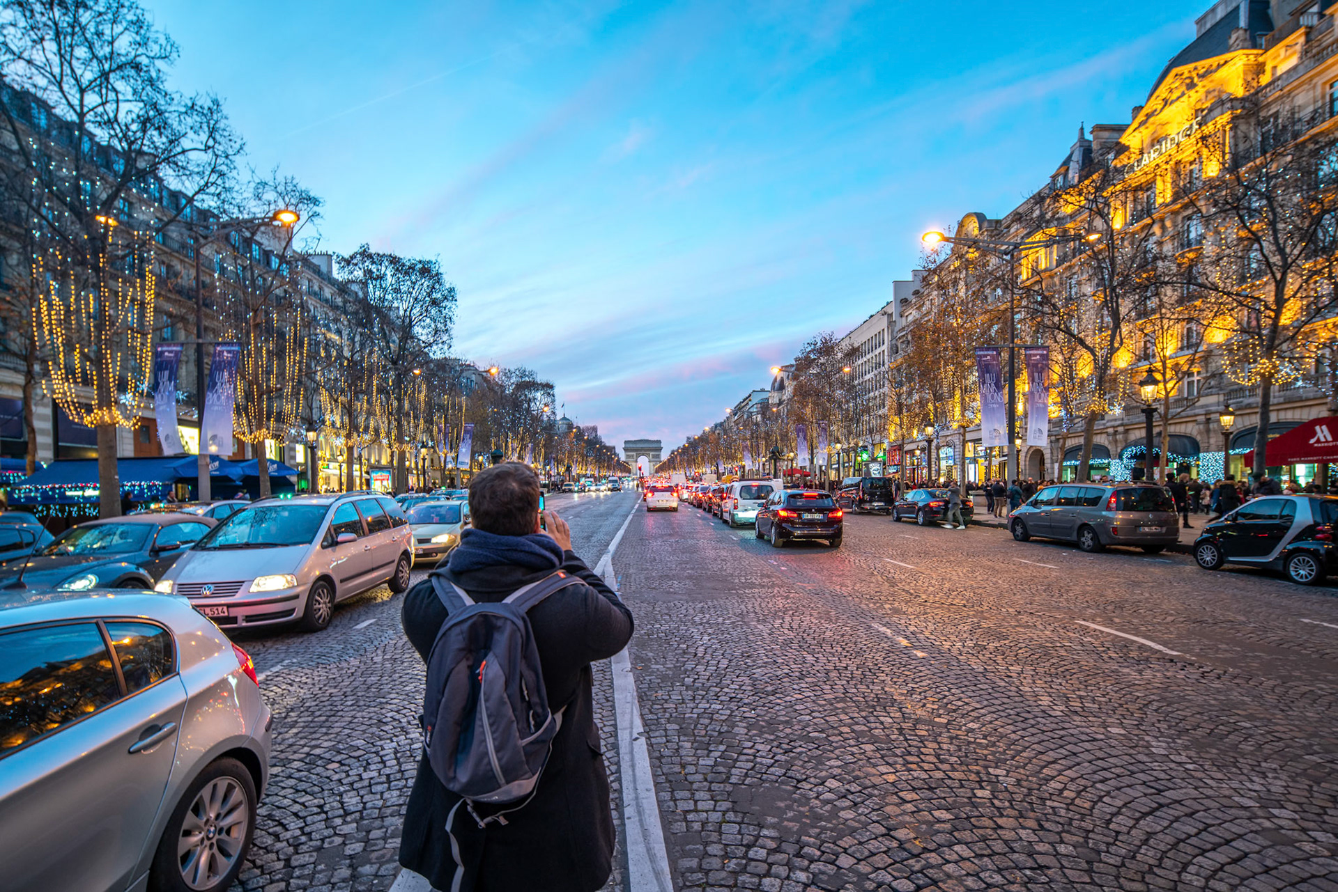 A tourist photographs the festive illumination along the Champs-Élysées at dusk.