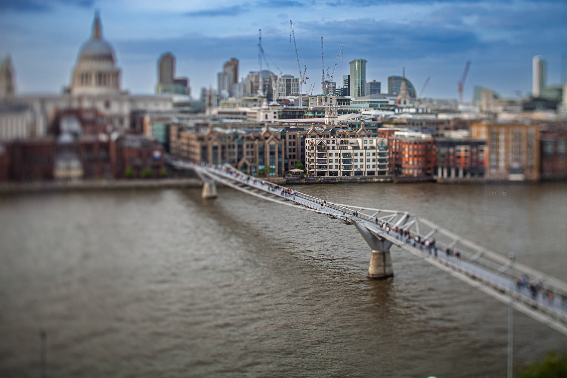 People walk across Millennium Bridge over the River Thames with St Paul Cathedral in the background. Cityscape of London is visible.