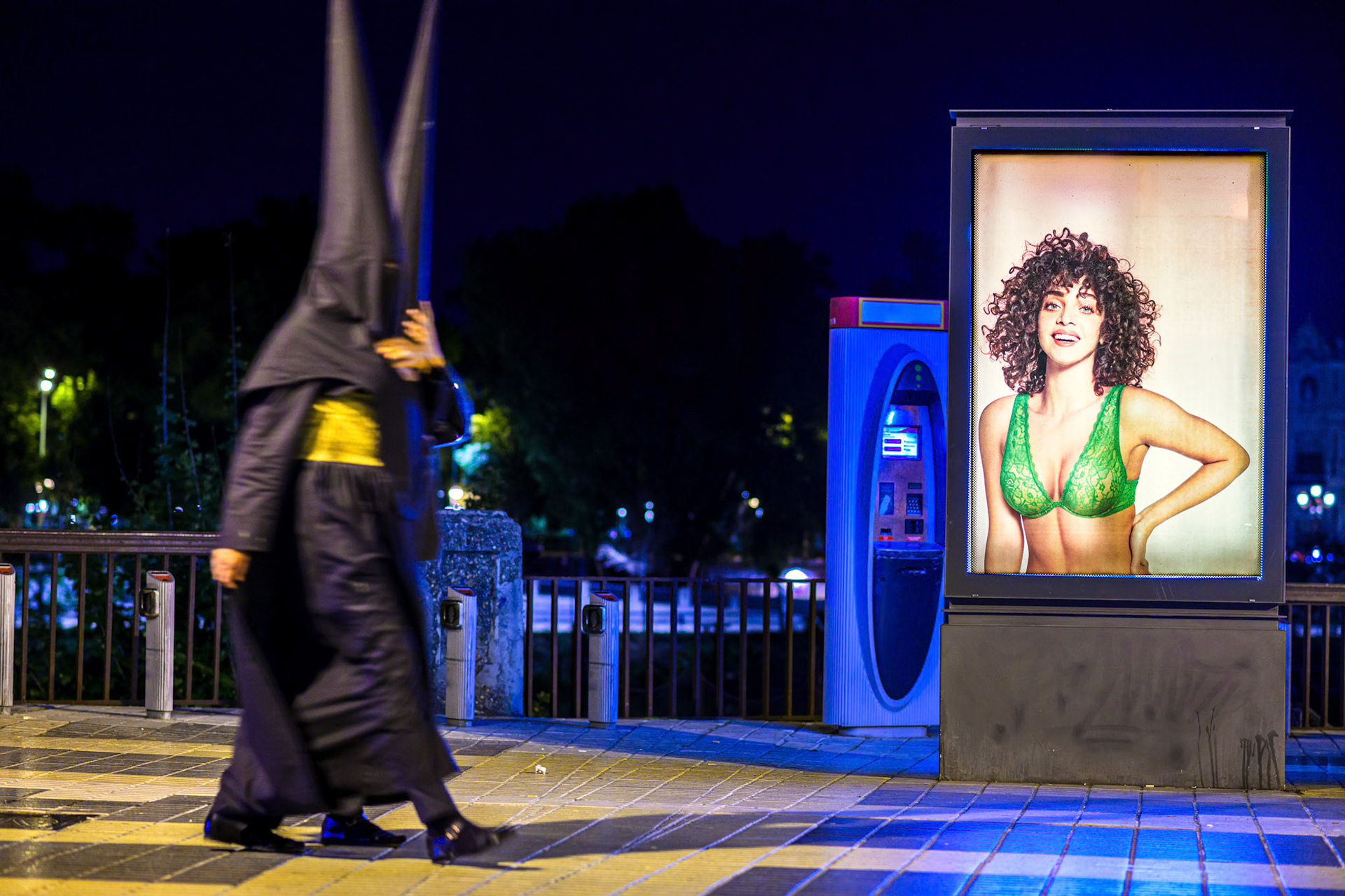 Two black-robed Holy Week penitents walk past an illuminated advertising screen displaying female lingerie at night in Seville, Andalusia, Spain.