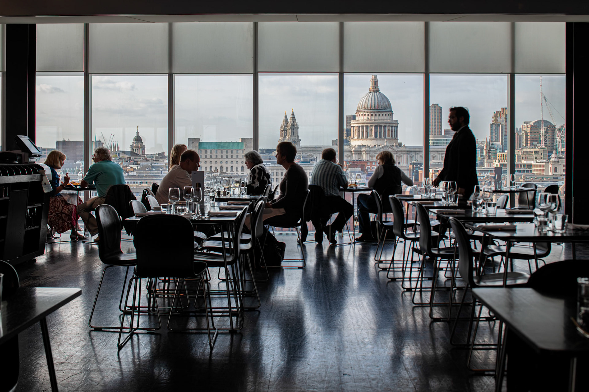 People enjoy their meals and drinks at the Tate Modern restaurant while St Paul Cathedral is visible through the large windows in the background.