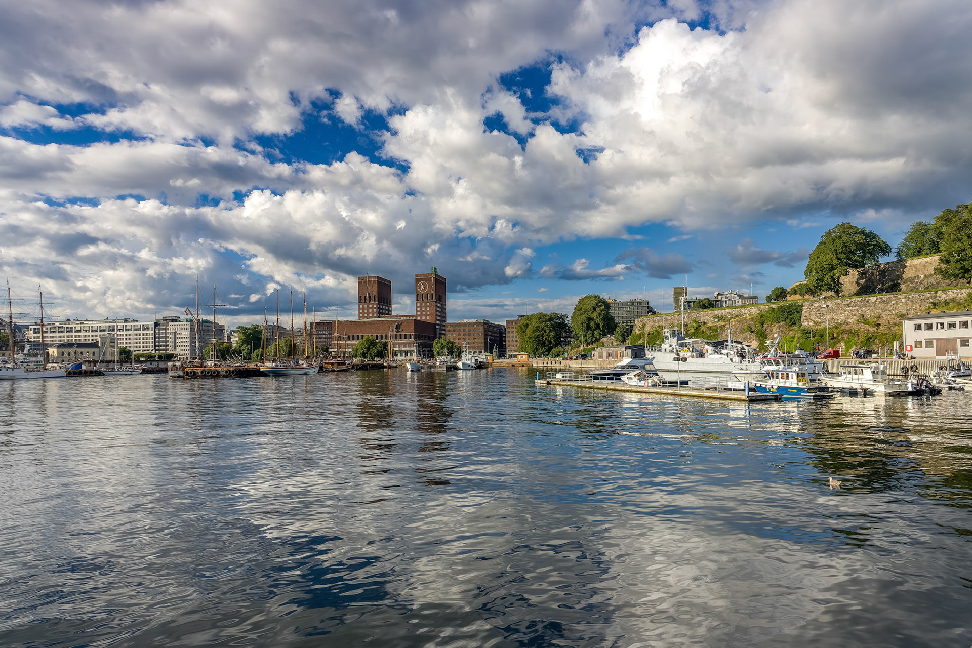 Oslo City Hall stands majestically beside the calm waters of the Fjord under a backdrop of fluffy clouds and blue sky.