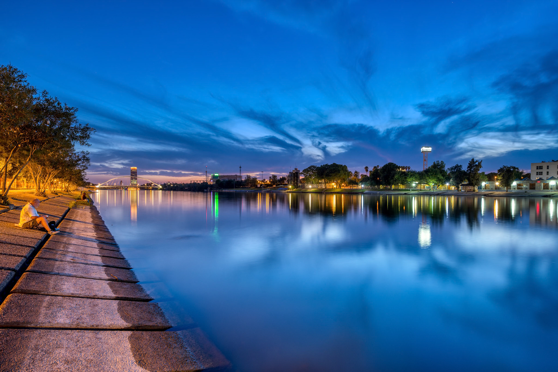 Sunset colors reflect on Guadalquivir River, showcasing La Cartuja Island, Barqueta Bridge, and Torre Sevilla in serene evening light.