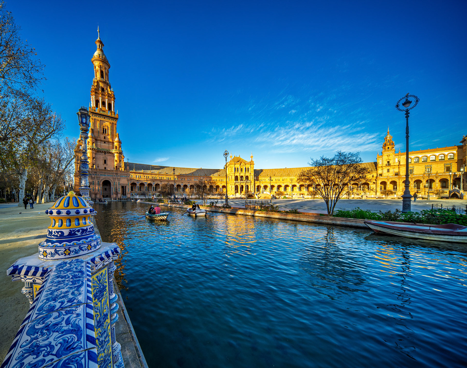 The north tower of Plaza de Espana rises beautifully above the canal, bathed in golden light as boats float lazily on the water at sunset.