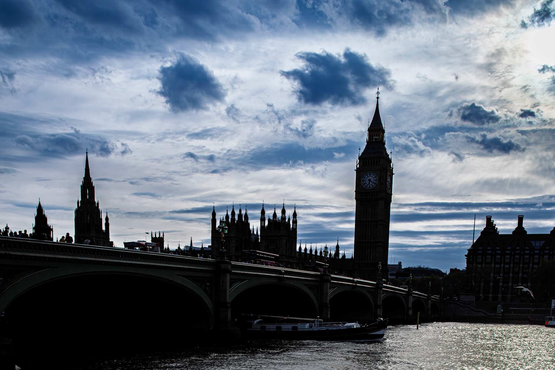 Big Ben stands tall against the dark clouds over the Thames River in London. The bridge leads to historic buildings along the water.