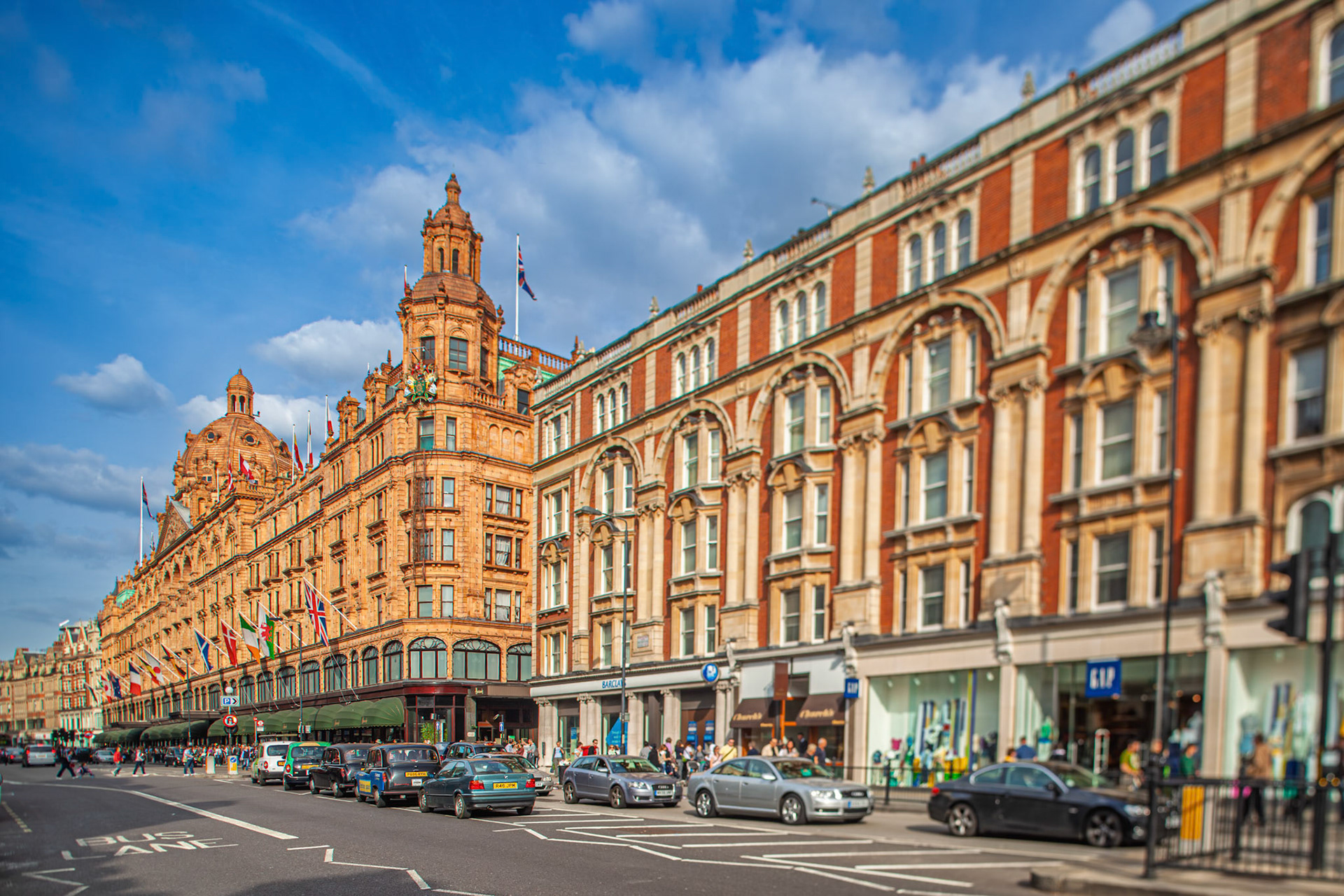 The Harrods building stands tall on Brompton Road in London, with people walking and cars driving nearby under a clear sky.