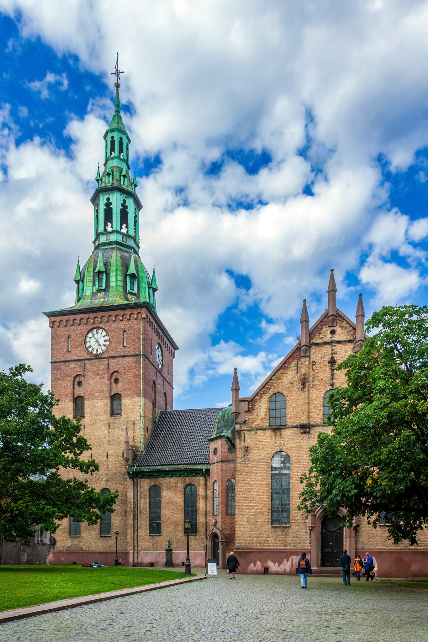 Visitors admire the striking bell tower of Oslo's cathedral under a bright blue sky, surrounded by historic buildings and greenery.