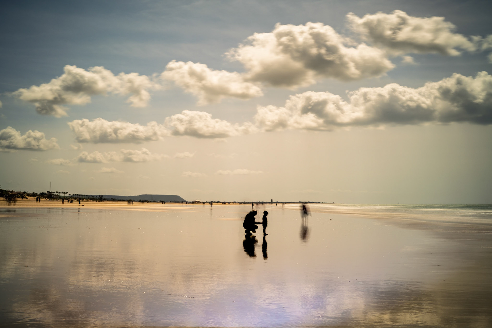 A child walks with his father on Conil's winter beach, creating reflections on the wet sand while ghostly figures drift in the moody atmosphere.