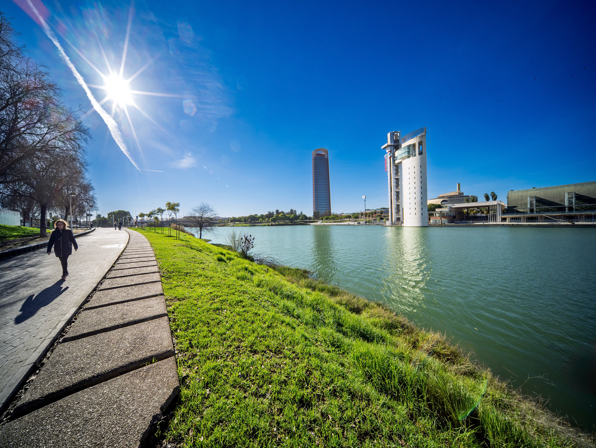 Seville, Spain, Dec 29 2021, Walkers enjoy a sunny day along the riverside path in Seville, framed by green lawns and shimmering water reflections by Torre Sevilla and Torre Schindler.
