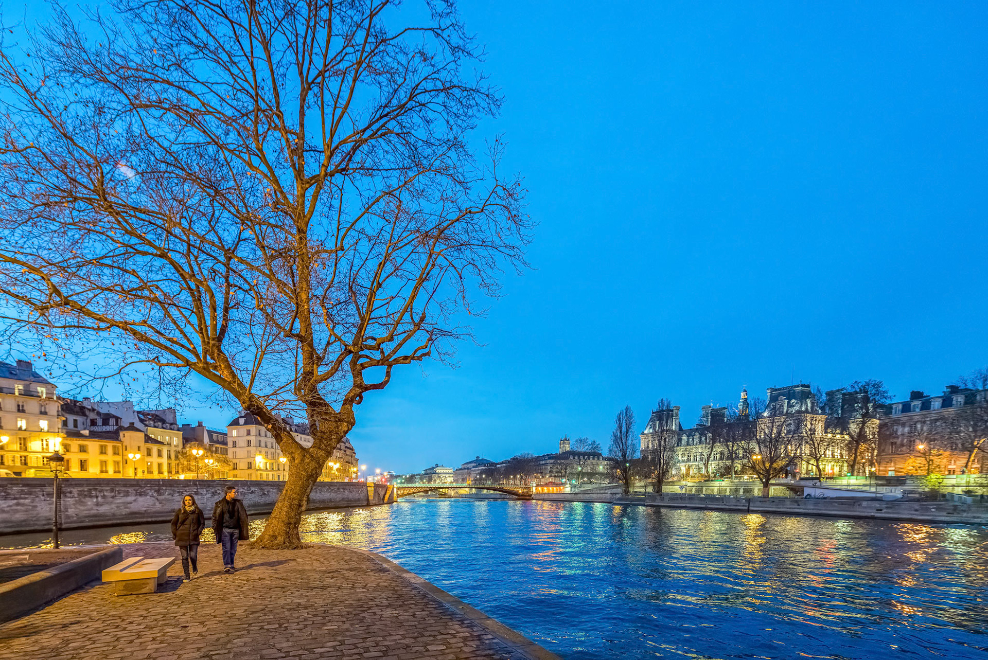A couple enjoys a twilight walk along the Seine River, with the ambient glow of Paris around them.