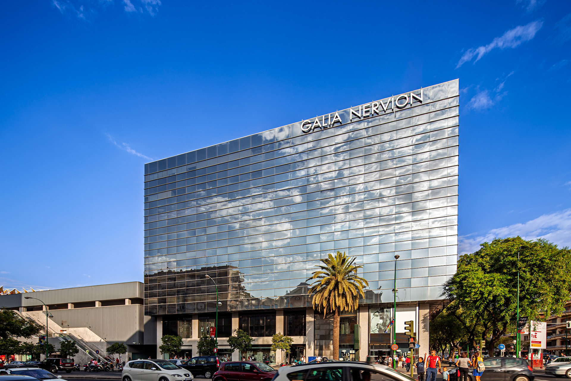 See Galia Nervion Building, a modern glass structure in Seville, Spain. Captures urban architecture and city life in a bright, sunny daytime shot.