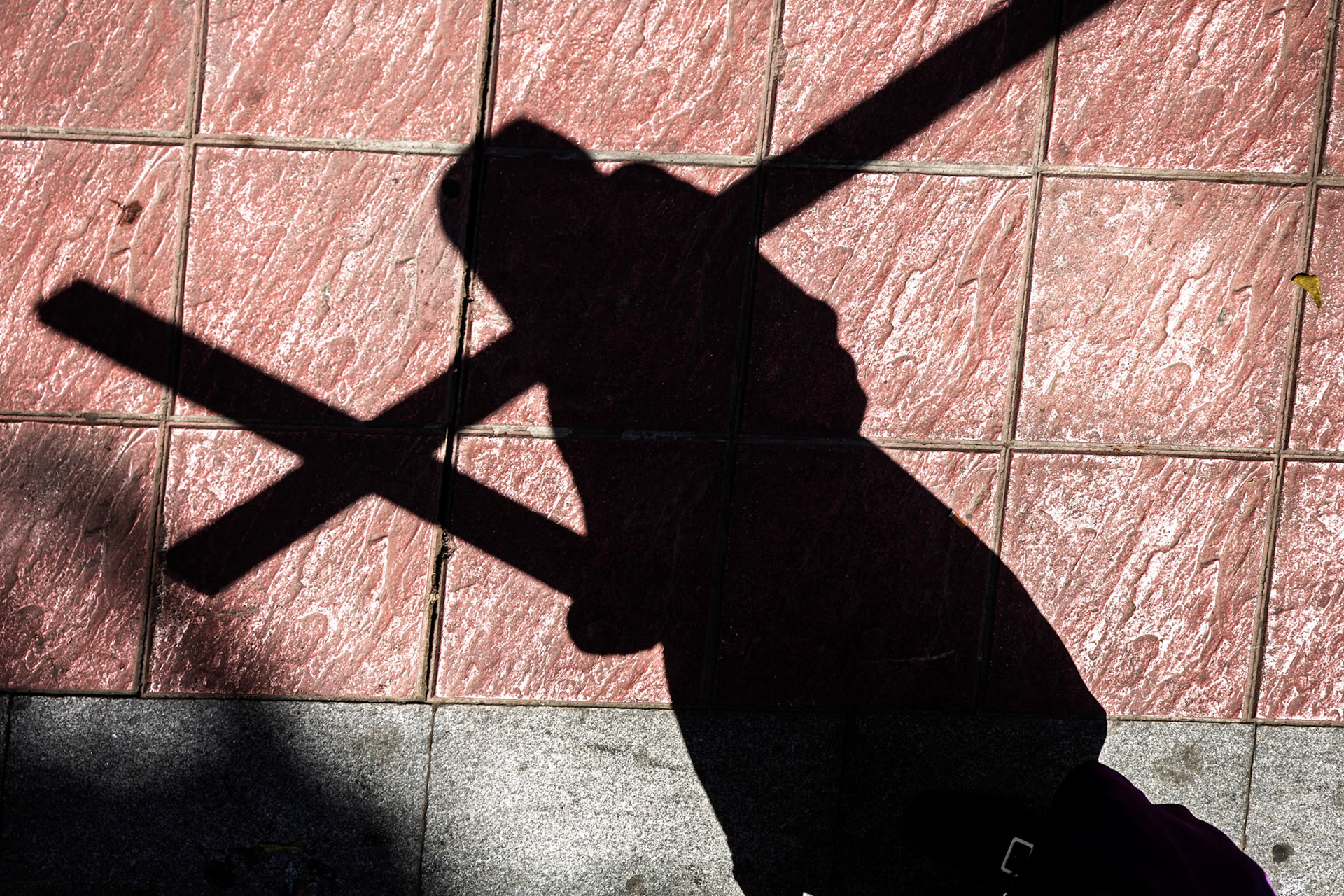 A hooded penitent walks with a wooden cross, casting a dramatic shadow on the pavement during Holy Week celebrations in Seville.
