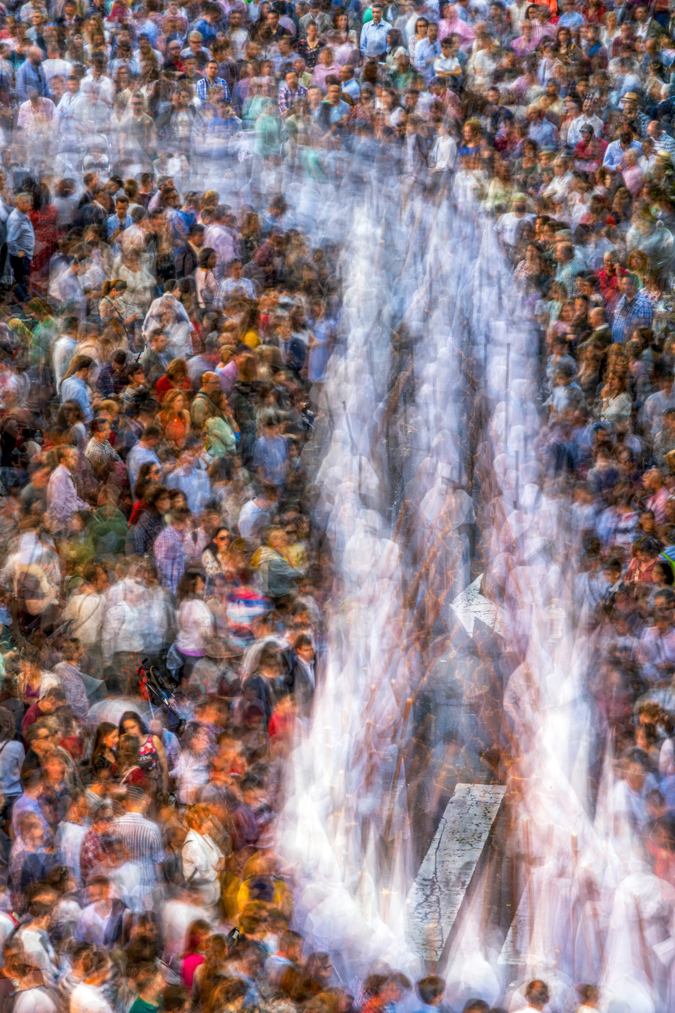 A vibrant, slow exposure captures white-robed Nazarene penitents amidst a dense crowd during a Holy Week procession in Seville, Andalusia, Spain.