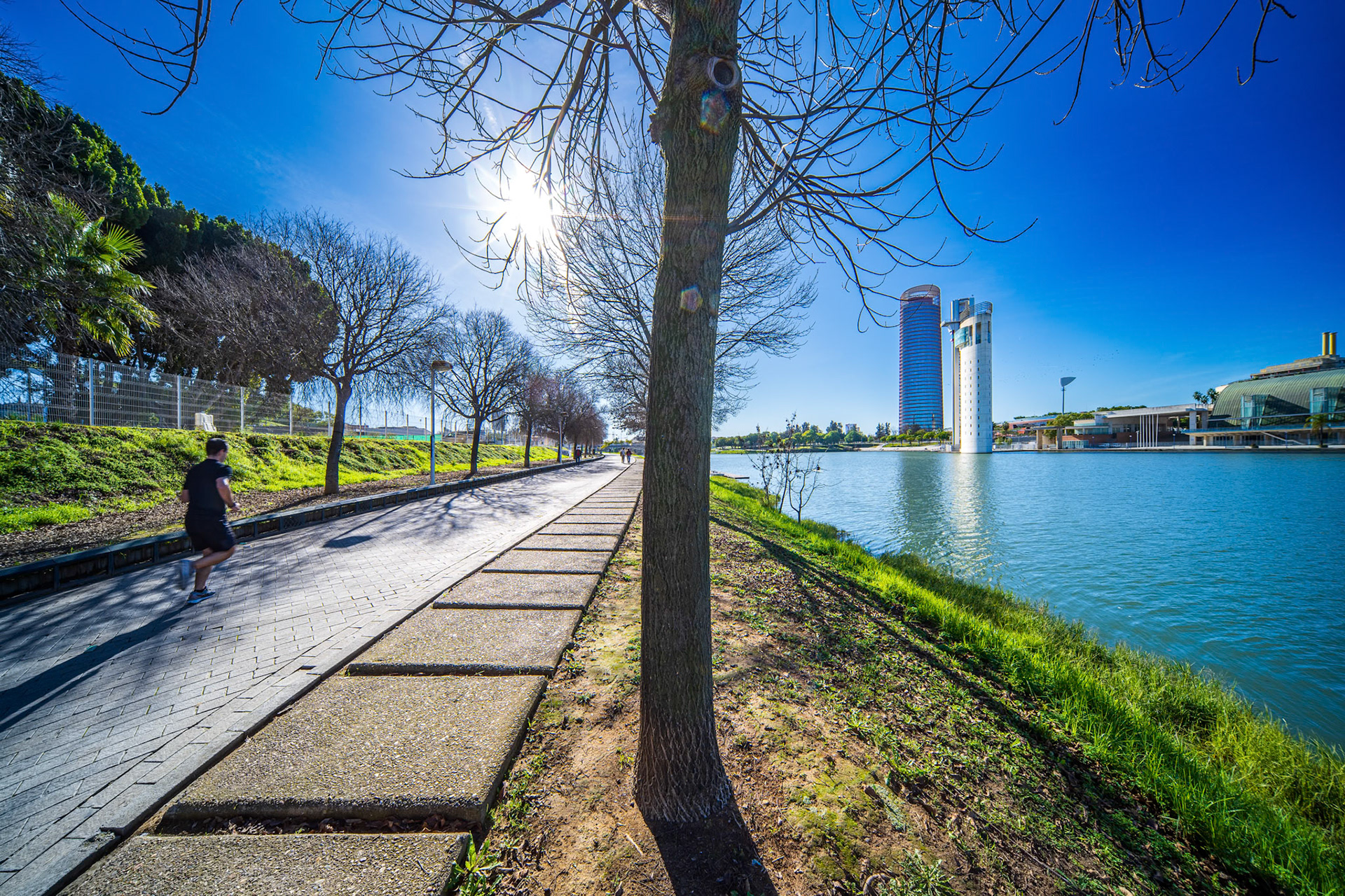 Runners pace the riverside path in Seville on a sunny winter day, with Torre Sevilla and Torre Schindler rising beyond the calm water.