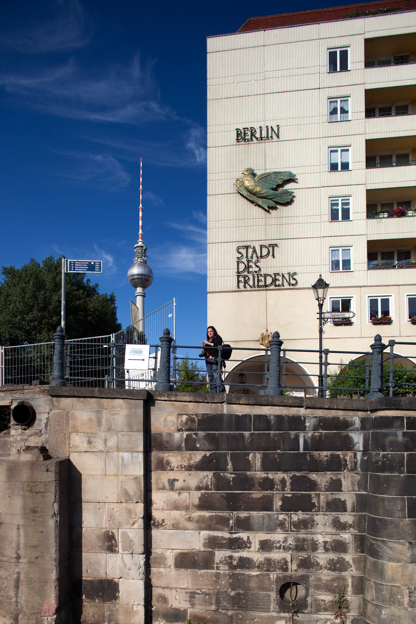 A historic GDR sign is displayed on a building by the Spree river, with the Fernsehturm visible in the background.