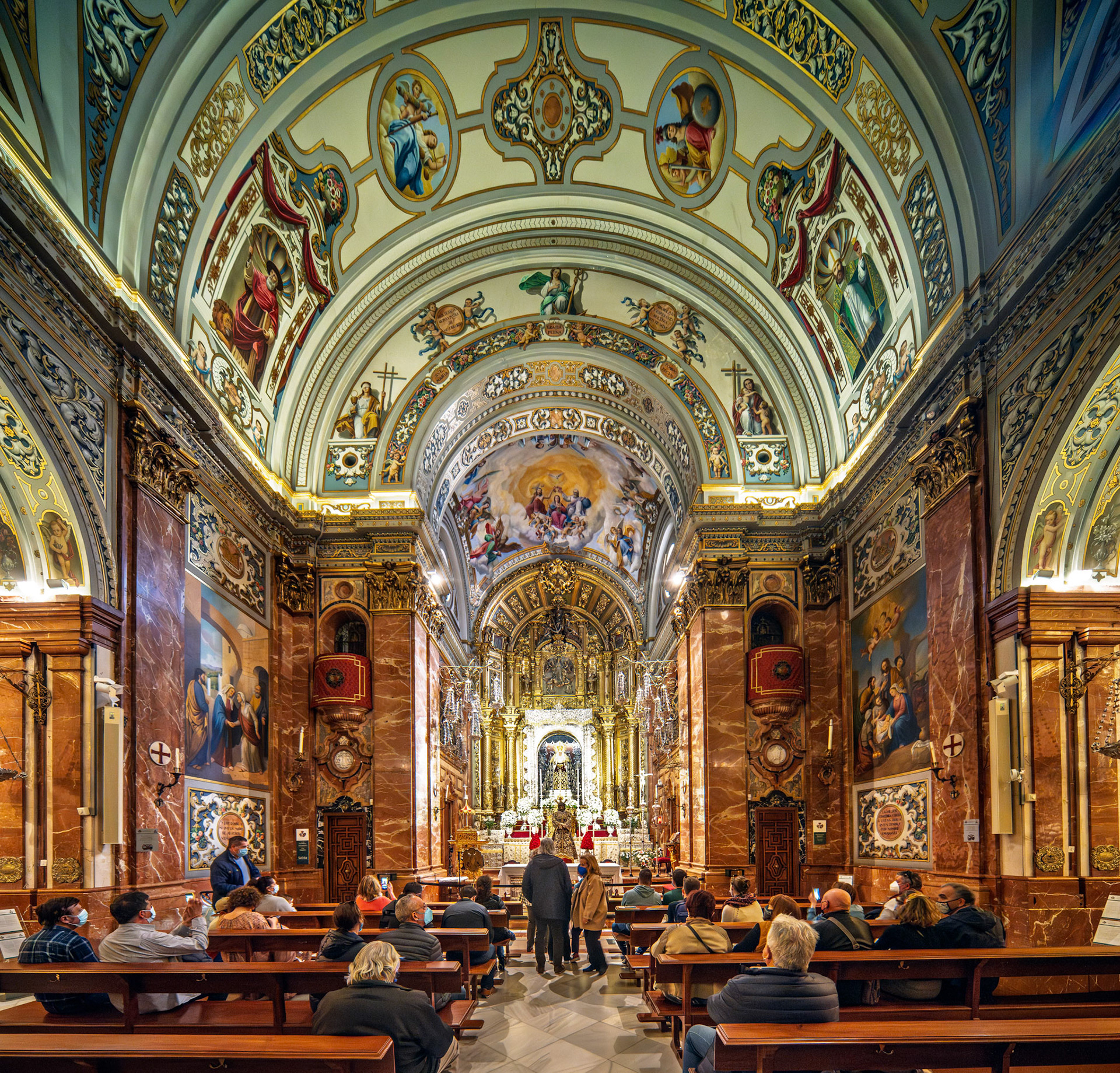 Visitors admire the stunning interior of Basílica de la Macarena, featuring gilded vaults and harmonious pew alignment beneath the altar.
