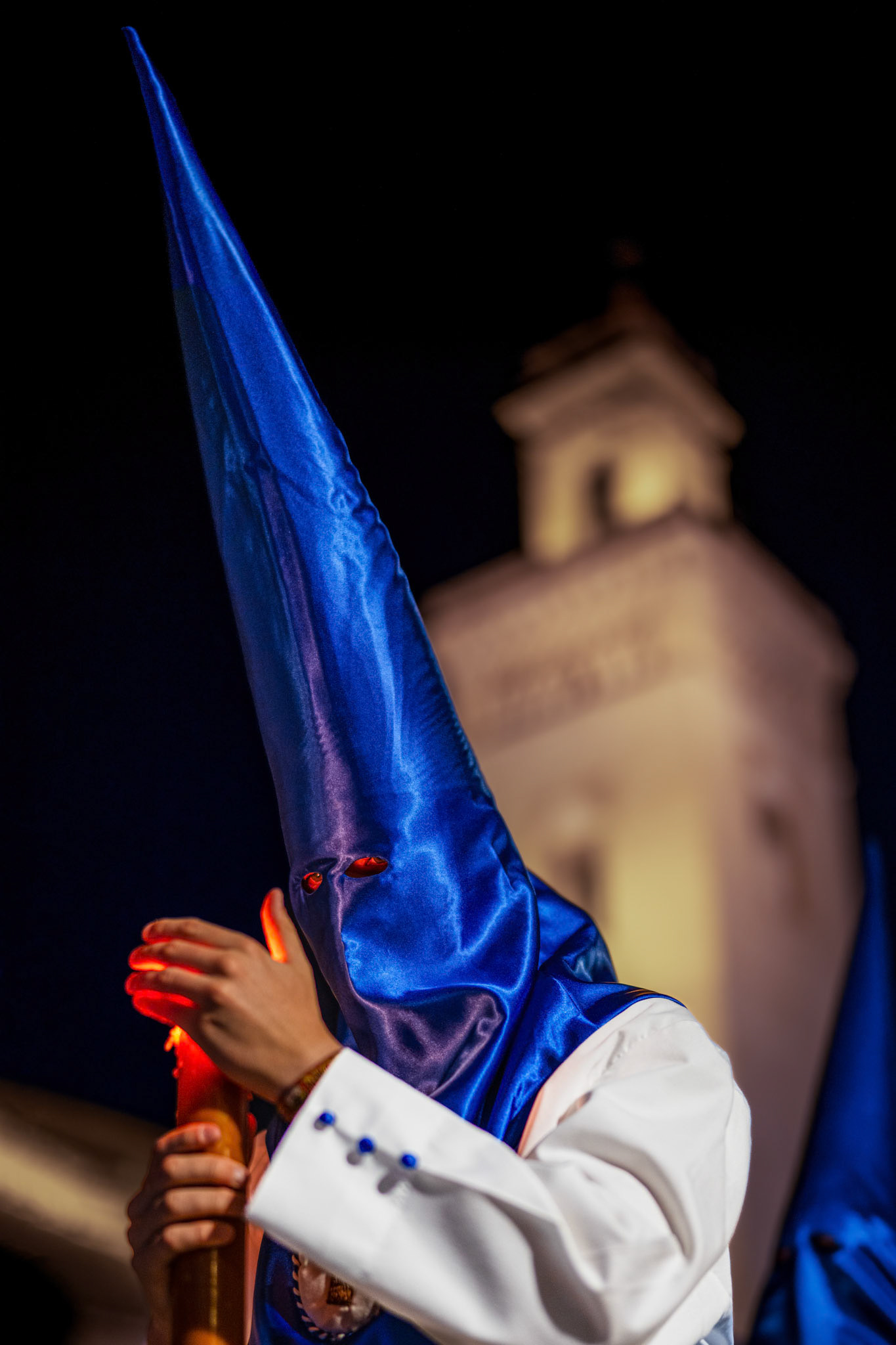 A Nazarene of the Hiniesta Brotherhood protects his processional candle flame on Palm Sunday night in Seville, Spain, with the San Marcos Tower in the background.