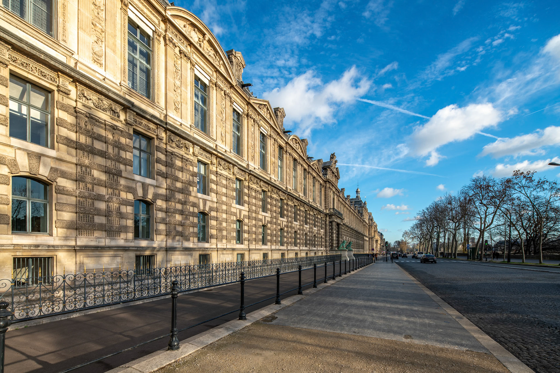 The Louvres facade basks in sunlight with blue skies and a tree-lined path.