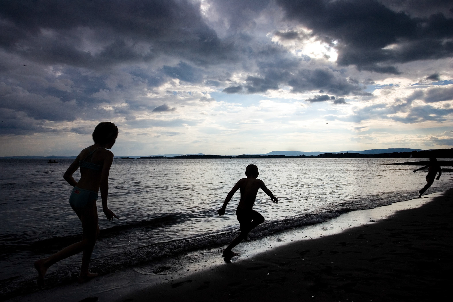 Silhouetted children run along the shoreline at Paradise beach, enjoying a playful moment as the waves crash gently in Oslo.