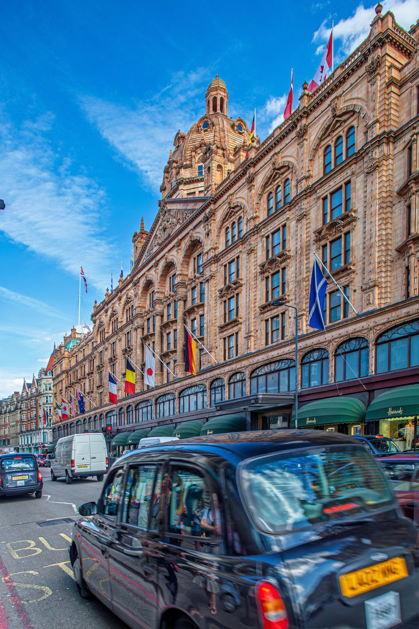 People walk along Brompton Road with the Harrods building in the background. Cars drive past on a busy day in London, England.