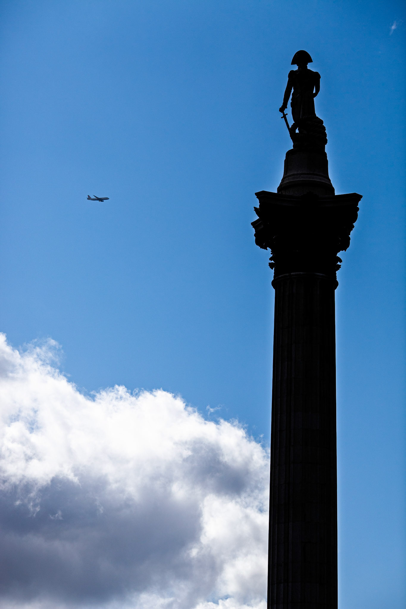 Nelson's Column rises in Trafalgar Square as an airplane flies through the clear sky, surrounded by clouds. Visitors gather below to admire the view.