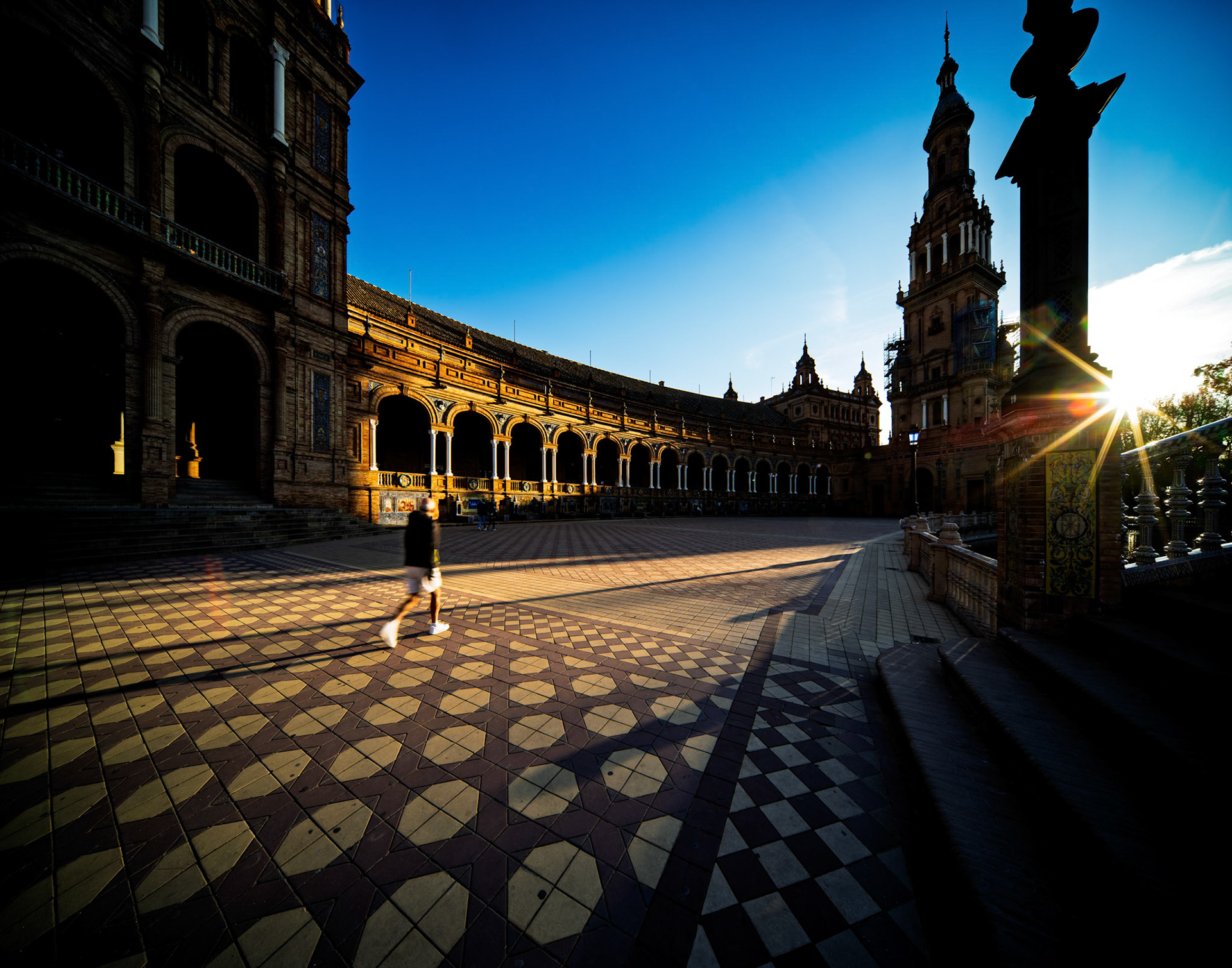 The wide esplanade of Plaza de España is bathed in golden light as a runner passes by, casting long shadows at dusk in Seville.