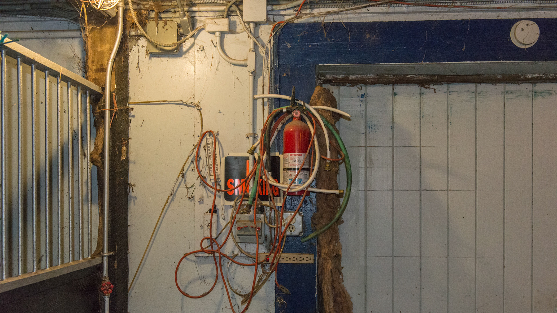 Located at the end of the barn aisle and next to the large barn doors was this. A fire extinguisher with access obstructed, a no smoking sign not visible, extension cords and a power strip, insulation stuffed into the door, I think the fly spray tubing is along the wall, and just plain filthy.