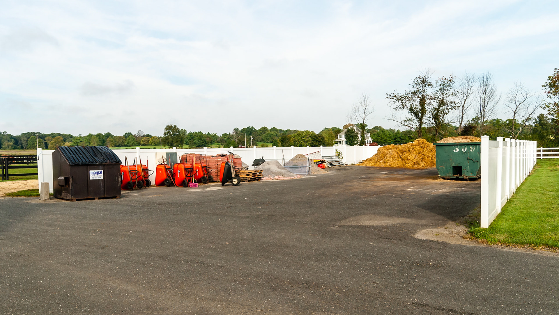 Shaving waste is piled into the roll off container.  Straw waste is piled on the asphalt.  Other supplies and equipment are organized and stored within this large walled off arera of this boarding barn.