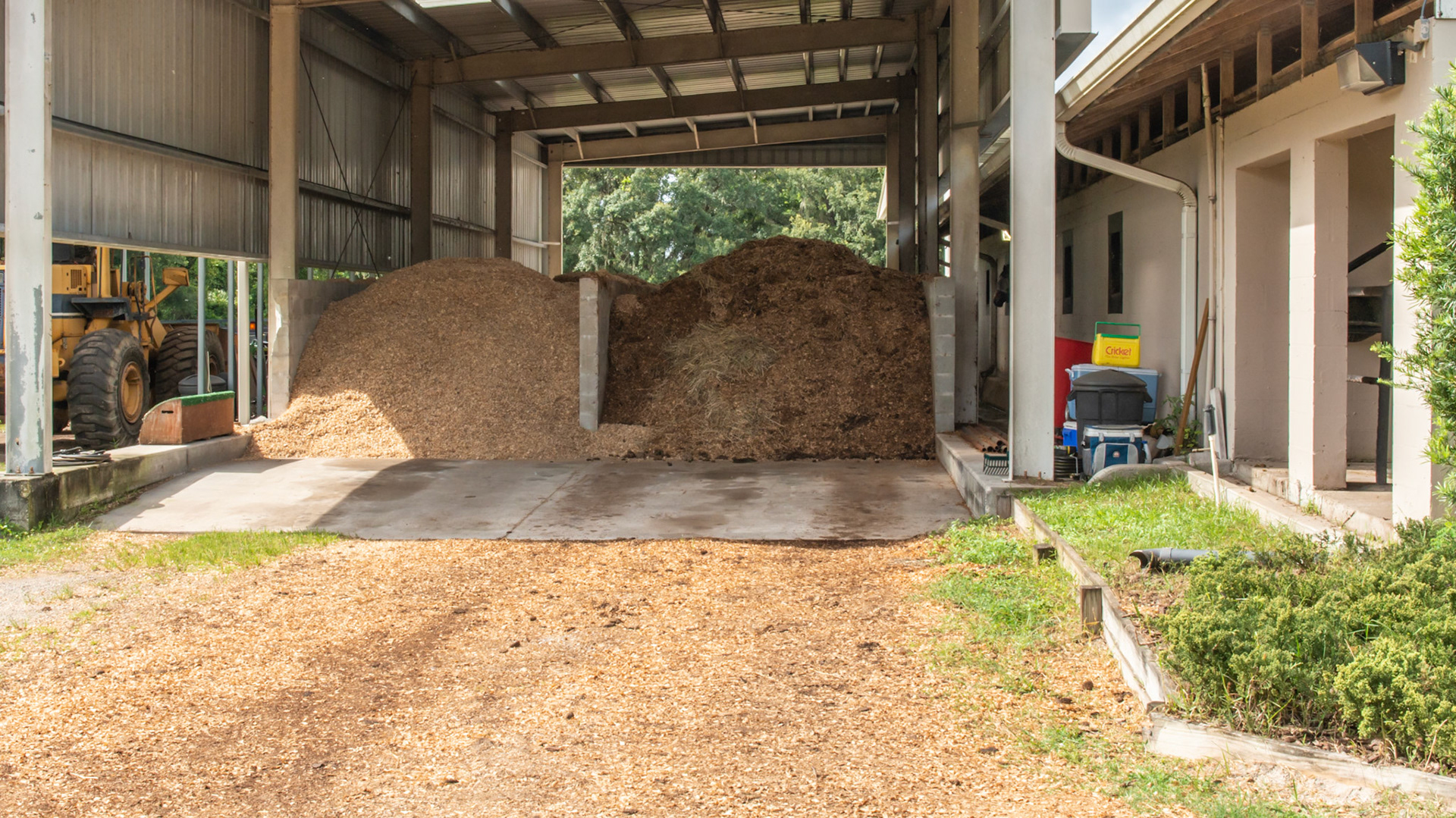 Covered bay for shavings and waste storage has a cement wall between them. The large yellow loader on the left manages this system. 1 of 3