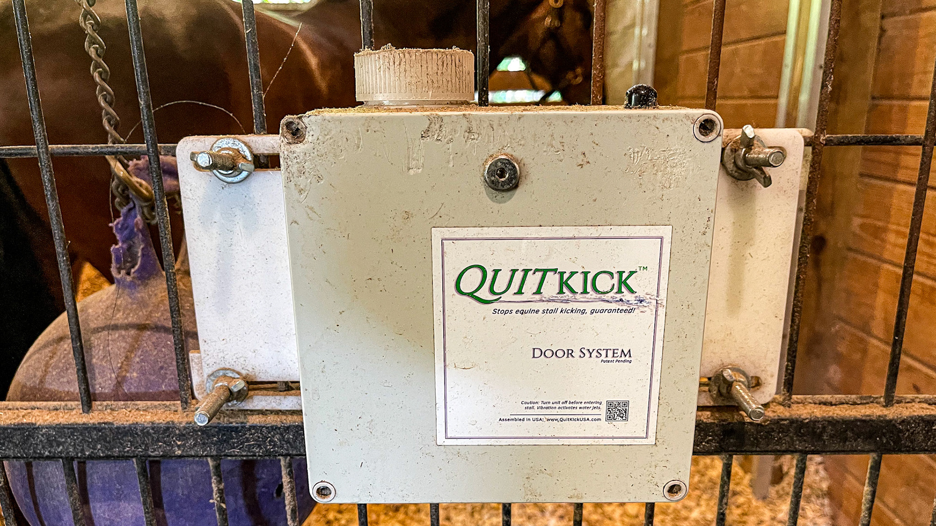 An anti-kicking device is attached to the stall door. When active, a kick triggers the release of a water spray towards the horse, suggesting to the horse to stop striking the door.