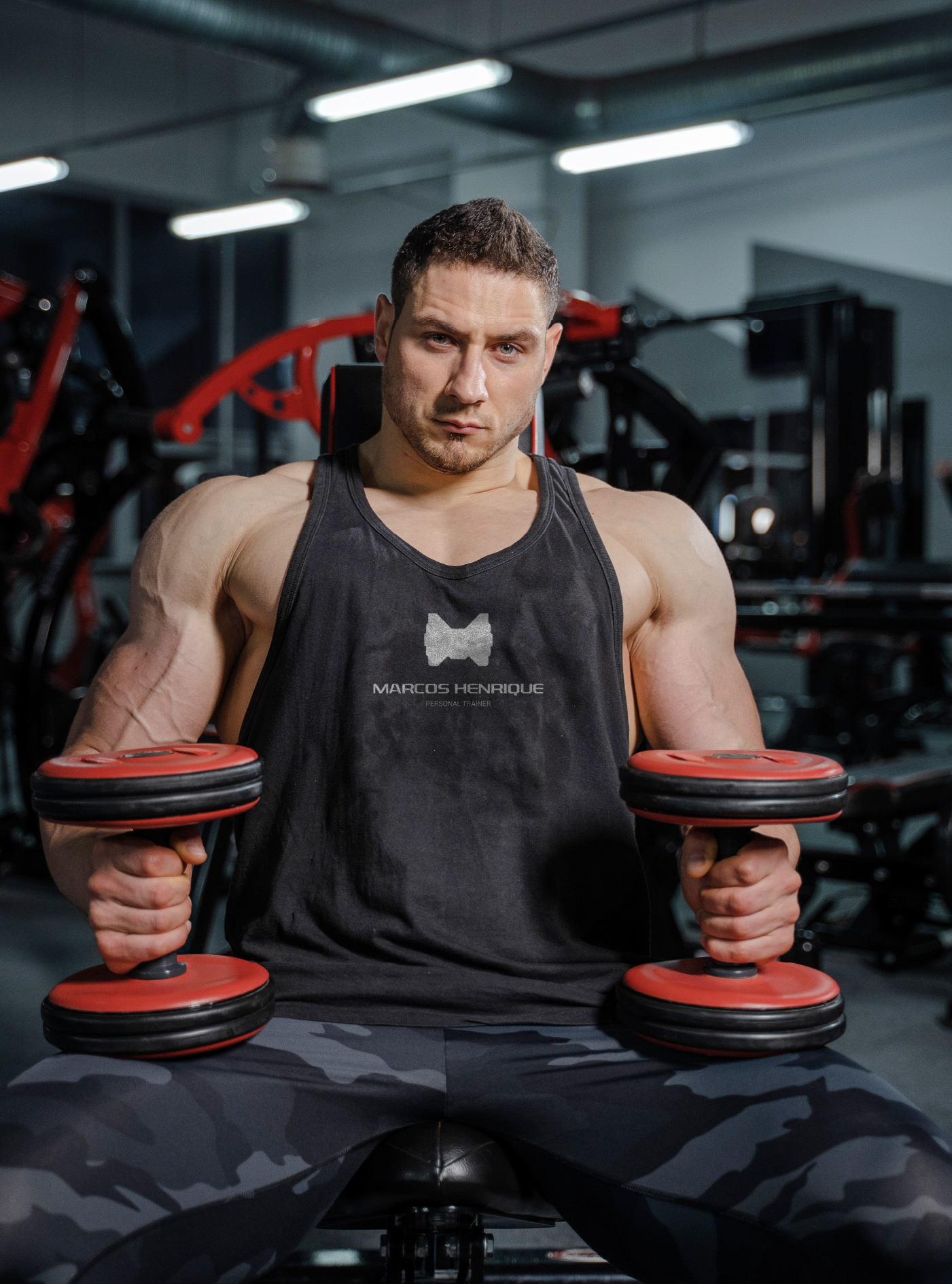 Homem musculoso em regata preta com logotipo da marca segurando halteres em academia com equipamentos vermelhos ao fundo