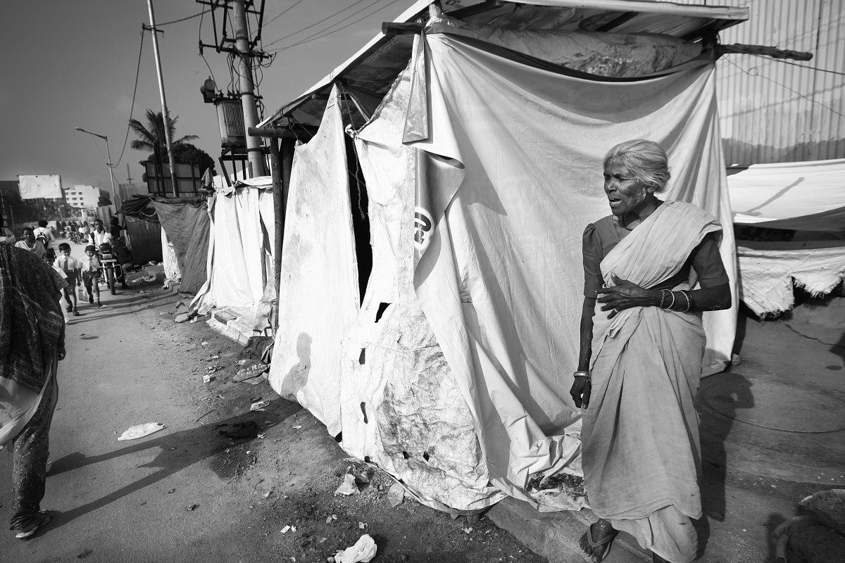An elderly woman wails at her condition as passersby on the road ignore her.