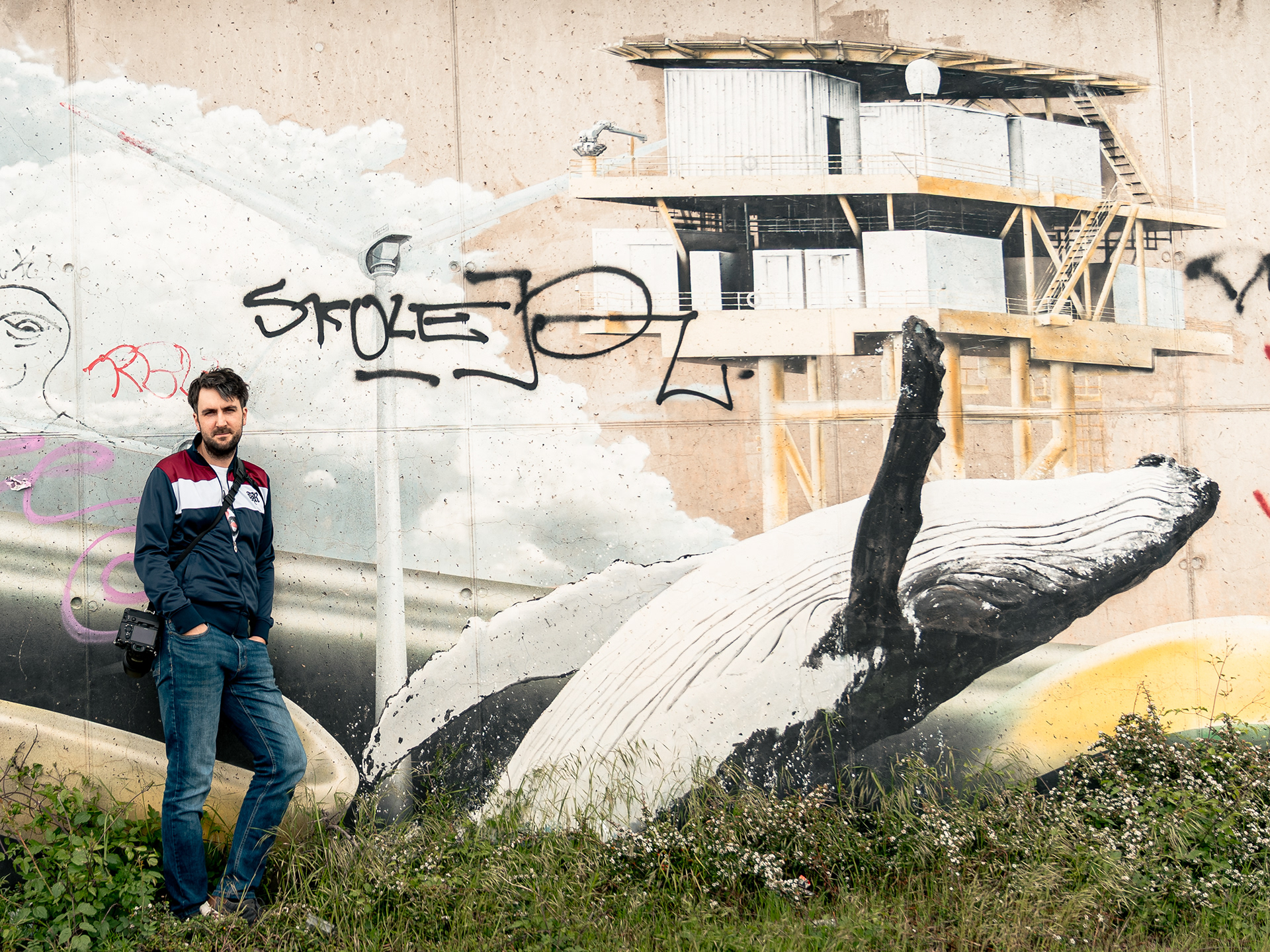 Der Fotograf und Gründer von SounDDcheck, Johannes Haupt, steht mit Kamera vor einer großflächigen Graffitiwand mit Wal-Motiv und industriellen Elementen. Das Portrait verbindet urbane Atmosphäre mit dokumentarischem Charakter und verweist auf die kreative Dresdner Kulturszene.
