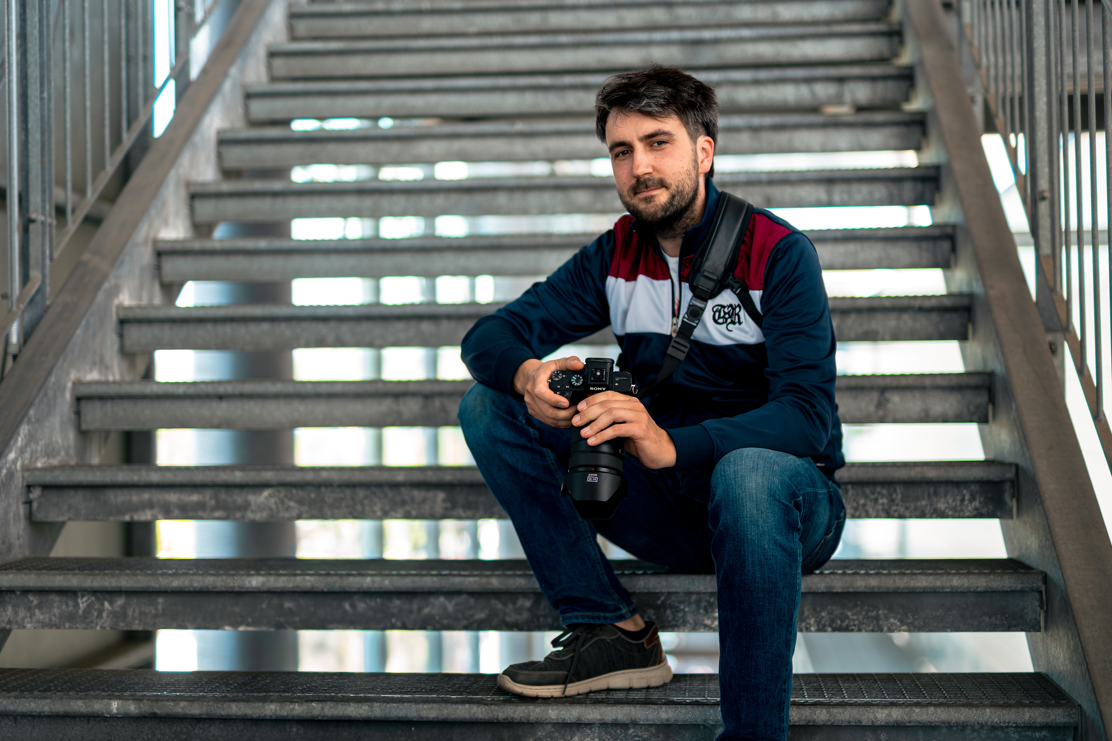 Der Fotograf und Gründer von SounDDcheck, Johannes Haupt, sitzt mit Kamera in der Hand auf einer Metalltreppe und blickt in die Kamera. Das Portrait zeigt ihn in urbaner Umgebung mit entspannter, dokumentarischer Atmosphäre.