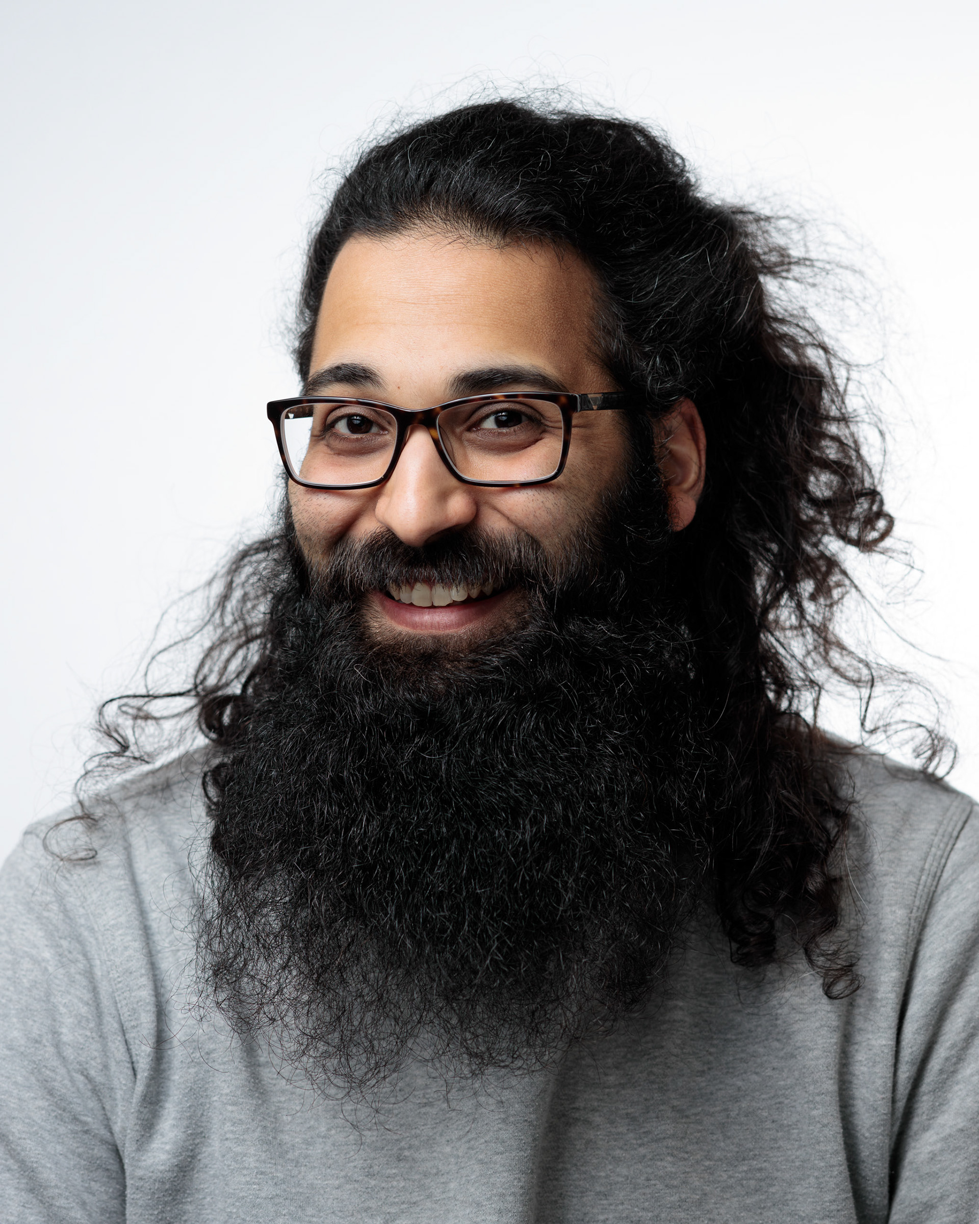Auf dem Bild ist ein Mann mit einem breiten Lächeln zu sehen. Er hat langes, lockiges Haar und einen vollen, buschigen Bart. Er trägt eine Brille mit einem dunklen Rahmen und einen grauen Pullover. Der Hintergrund ist weiß und unscharf, sodass der Fokus auf dem Mann liegt.