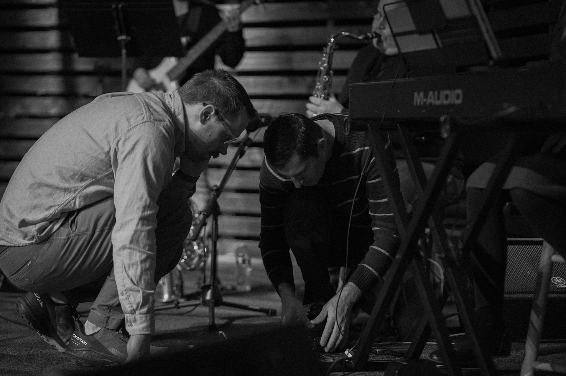 On Sunday morning at 7:06 AM, during rehearsal, David Iliyn (left), the sound guy, and Leo Foshka (right), a keyboard player, troubleshoot a sound issue with Leo's daughter, Sabrina Foshka's keyboard while the rest of the worship team waits.
