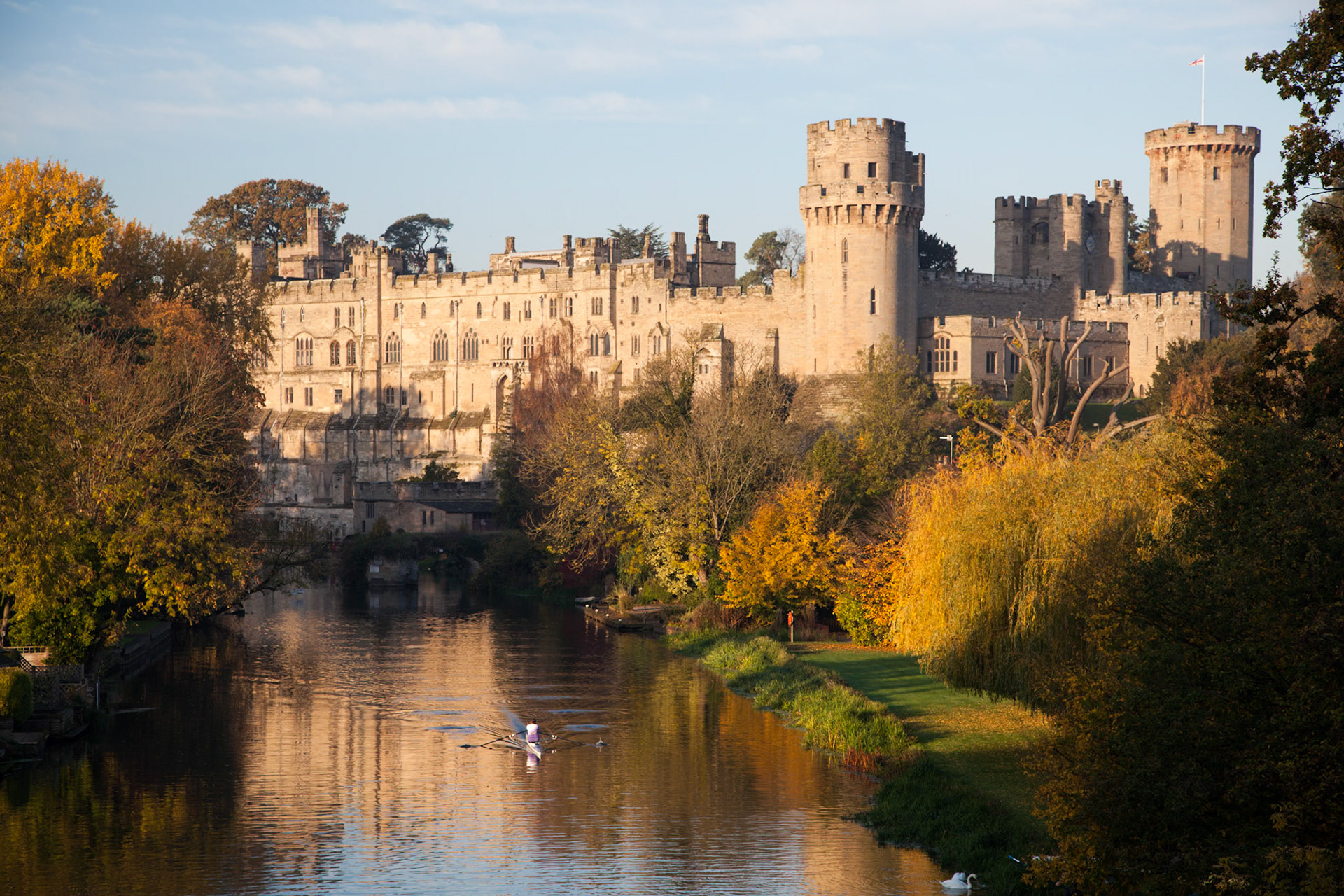 Warwick Castle reflected in the River Avon and lit by early sunlight with a rower sculling in the water