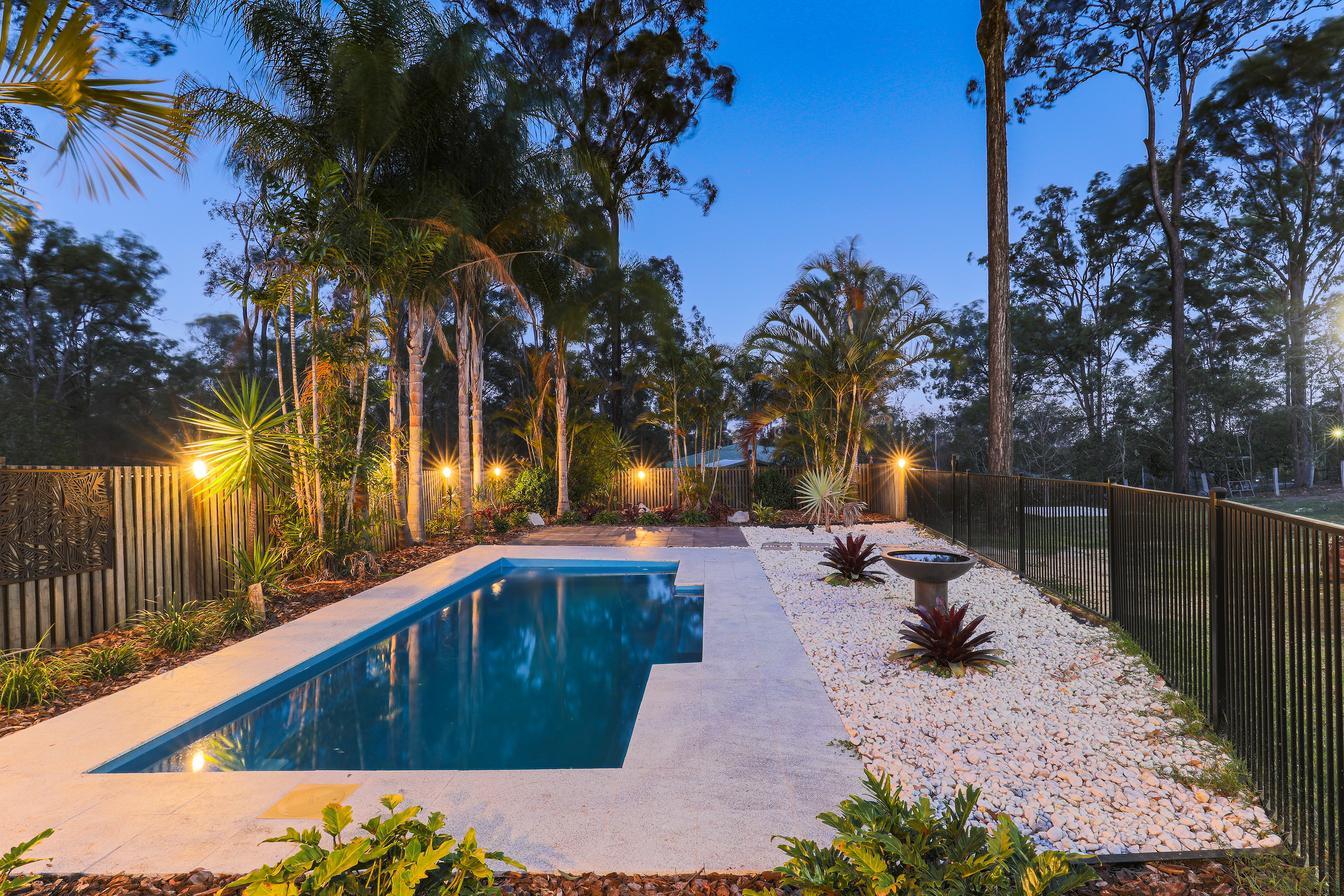ranquil backyard with a beautifully lit rectangular swimming pool surrounded by lush tropical landscaping, palm trees, and ambient evening lighting in Brisbane, QLD. Professional real estate photography by Jeremy Murphy.