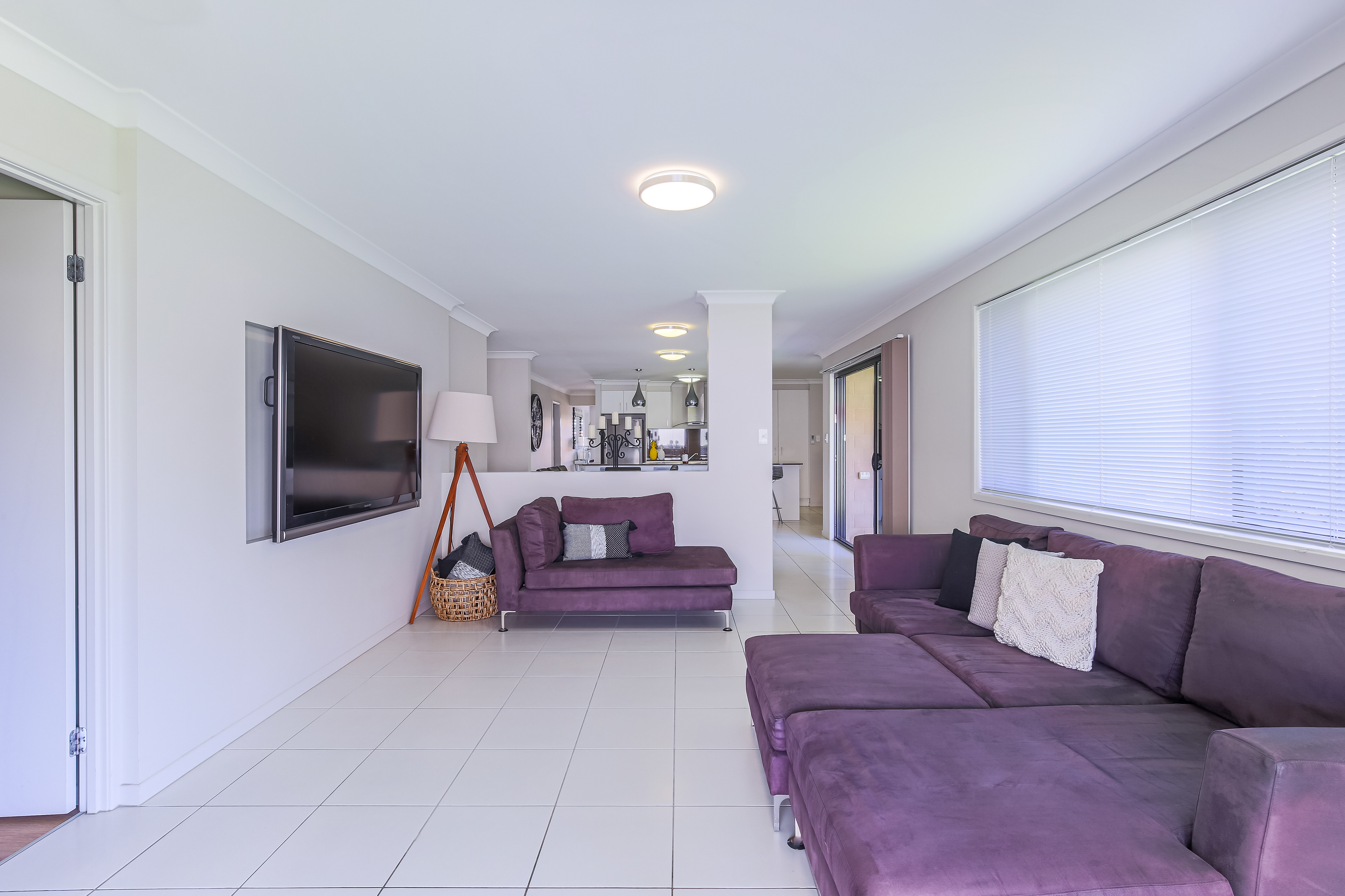 Modern living room with a purple sectional sofa, wall-mounted TV, and open layout leading to the kitchen, featuring large windows for ample natural light, located in Ipswich, QLD. Professional real estate photography by Jeremy Murphy.
