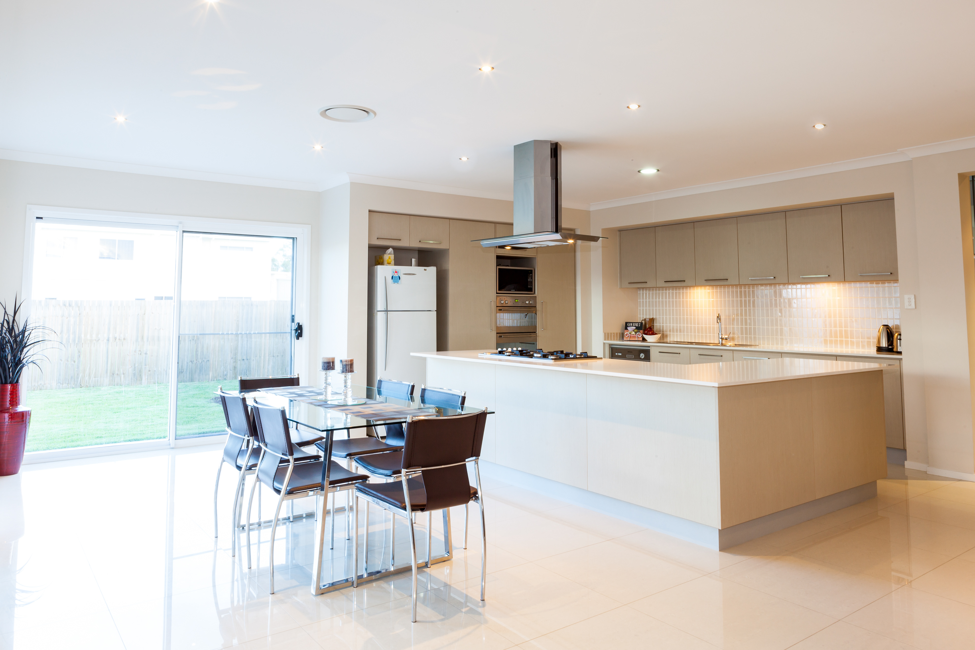 Spacious open-plan kitchen and dining area with modern appliances, large island, and glass dining table, featuring sliding doors that open to a backyard, located in Brisbane, QLD. Professional real estate photography by Jeremy Murphy.