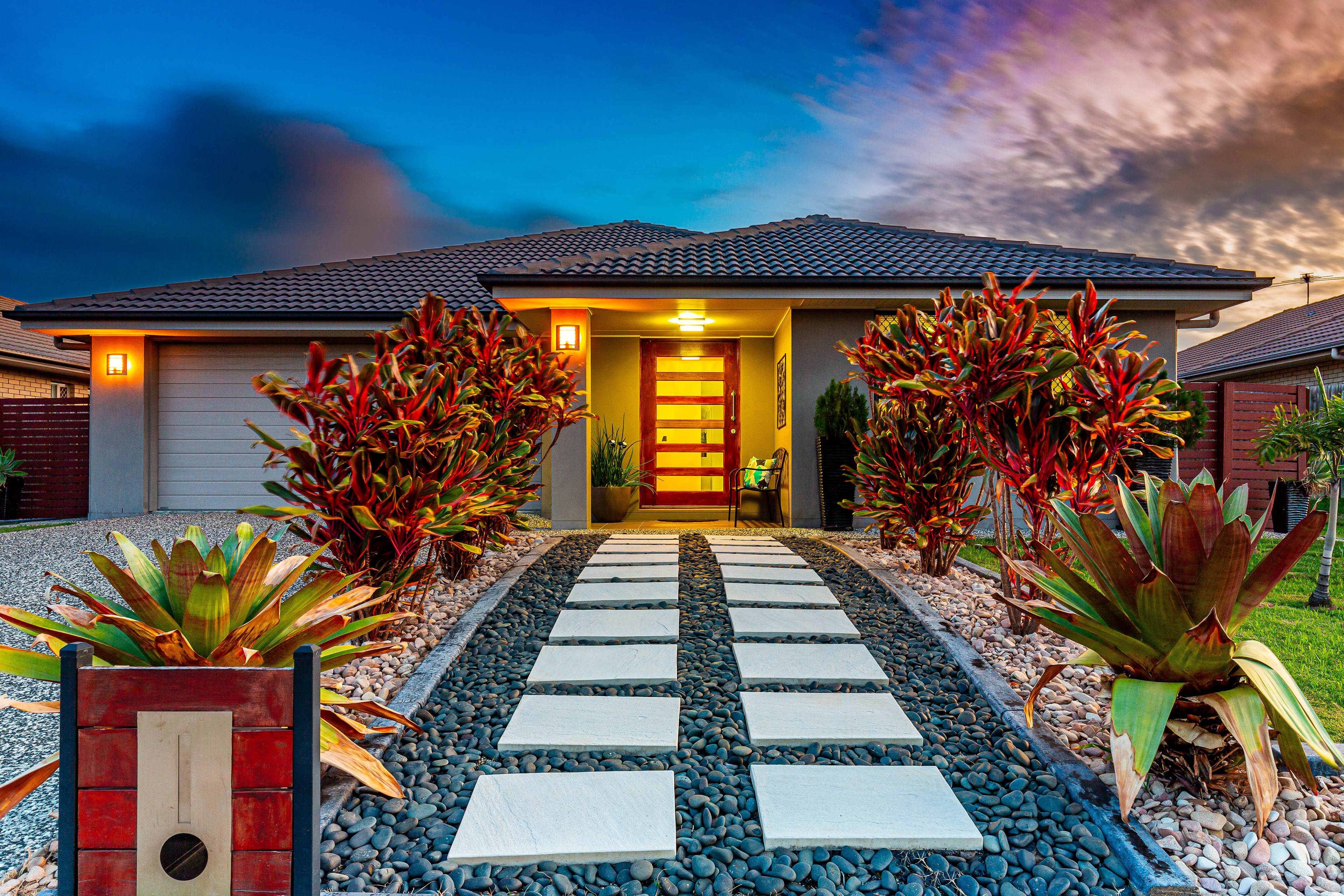 Stunning twilight real estate photograph of a modern Ipswich QLD home by Jeremy Murphy. The image showcases a beautifully lit pathway with sleek stepping stones, vibrant landscaping with red and green foliage, and a welcoming entrance featuring contemporary design elements. Perfect example of professional real estate photography in Ipswich.