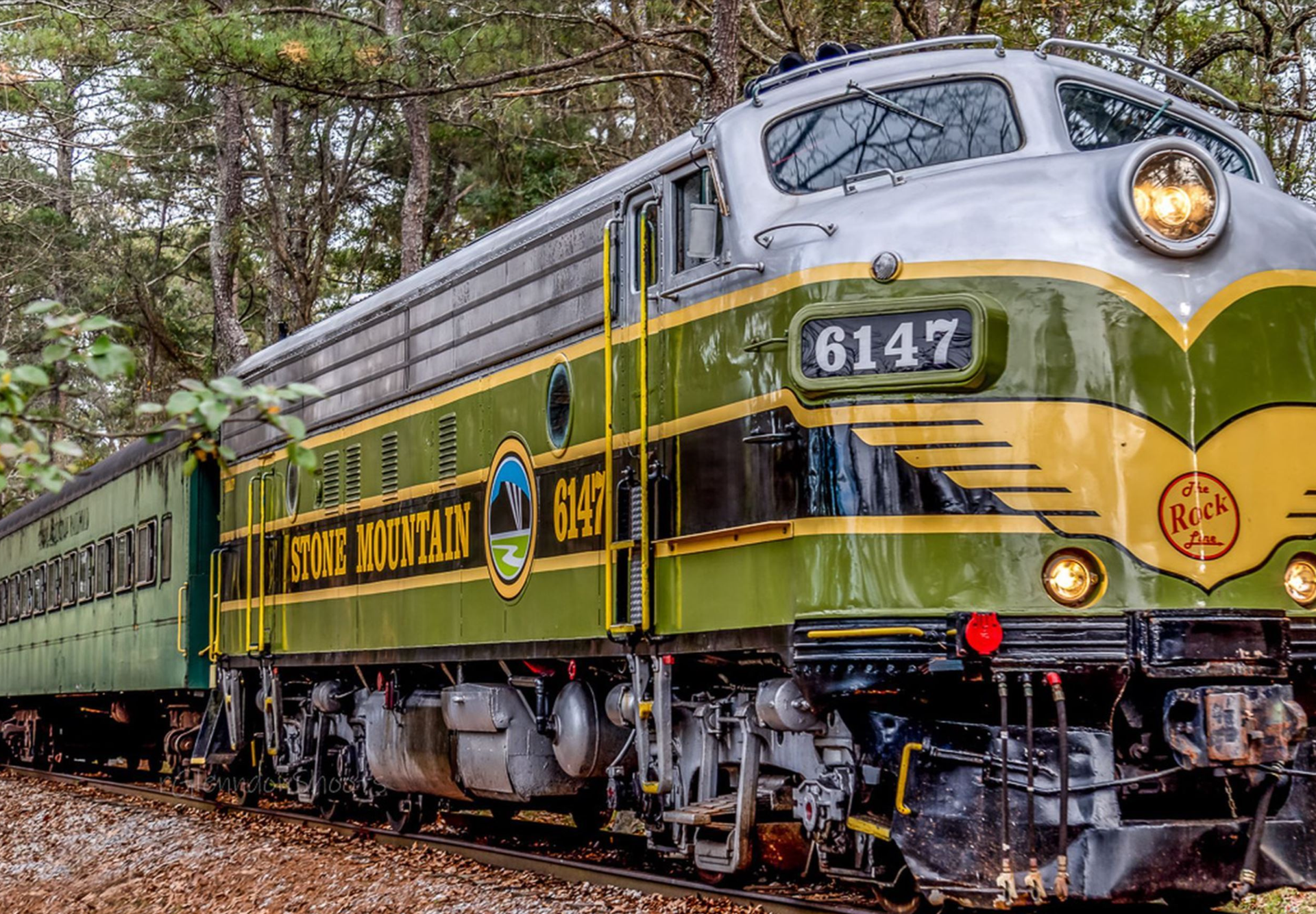 A green and yellow vintage diesel locomotive sits on the tracks under a partly cloudy sky, its front facing the viewer. The engine’s number “6147” is visible, along with red and circular emblems on the nose. Trees line the background and a person stands at a distance near the rail line.