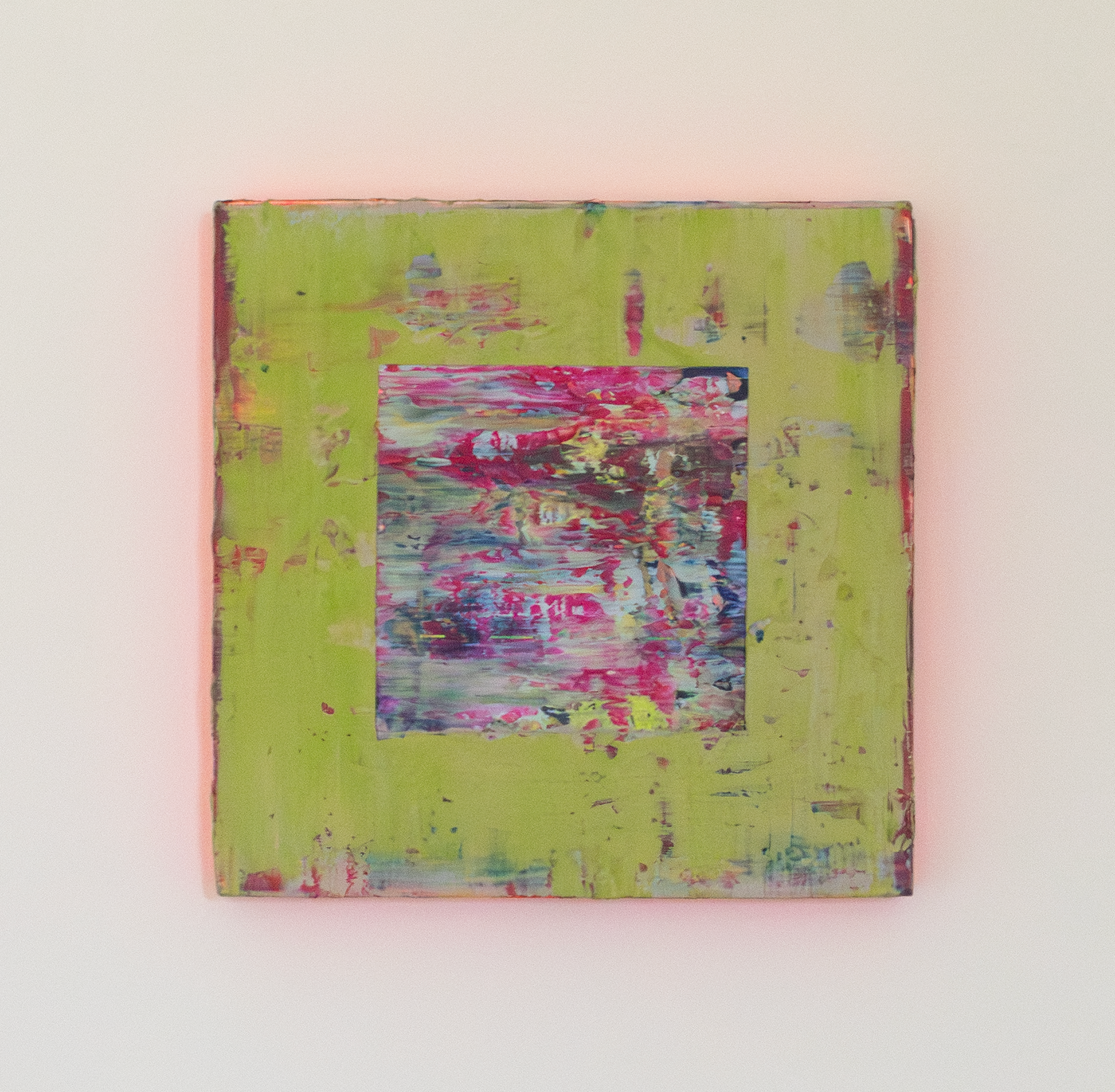 Front view of textural abstract painting with a smooth surface of layered colours that have been applied with a squeegee. The main colours are a soft green, with some blues and reds exposed in the thinner areas of green. In the centre there is a cut away square exposing a textural, layers painting underpainting with key colours of magenta, blue and greys. The edges of the painting are neon red which is reflecting on the white wall to make a red glow.