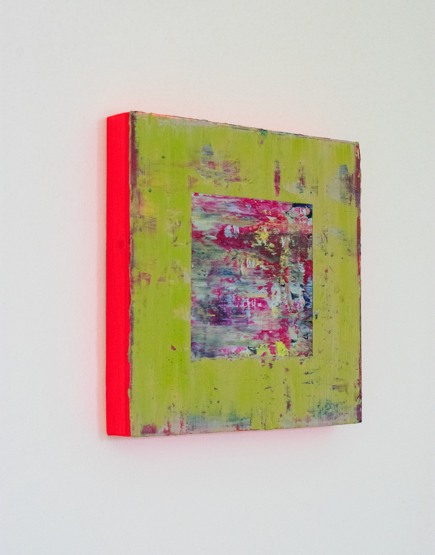 Left side view of textural abstract painting with a smooth surface of layered colours that have been applied with a squeegee. The main colours are a soft green, with some blues and reds exposed in the thinner areas of green. In the centre there is a cut away square exposing a textural, layers painting underpainting with key colours of magenta, blue and greys. The edges of the painting are neon red which is reflecting on the white wall to make a red glow.