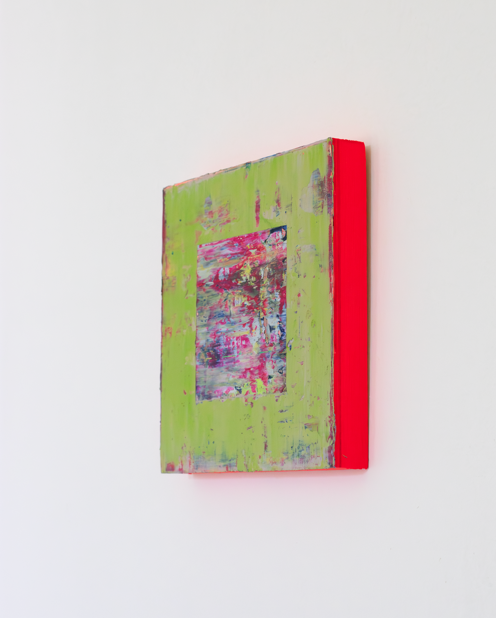 Right side view of textural abstract painting with a smooth surface of layered colours that have been applied with a squeegee. The main colours are a soft green, with some blues and reds exposed in the thinner areas of green. In the centre there is a cut away square exposing a textural, layers painting underpainting with key colours of magenta, blue and greys. The edges of the painting are neon red which is reflecting on the white wall to make a red glow.