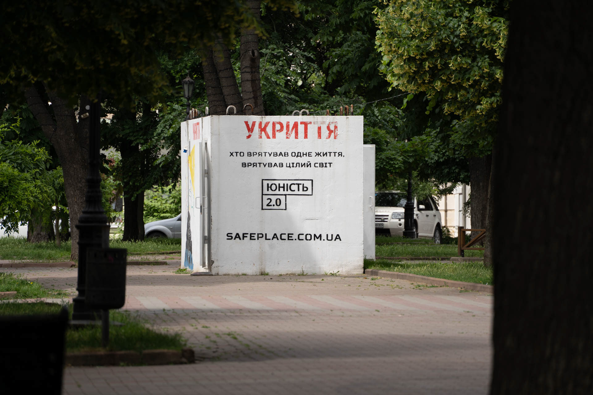 A public bomb shelter marked "Укриття" (Shelter) stands in a park in Odesa, Ukraine, on June 13, 2026. The structure carries the inscription "Хто врятував одне життя, врятував цілий світ" (Whoever saves one life saves the world entire), the branding "Юність 2.0" and the website safeplace.com.ua, with a car parked nearby.